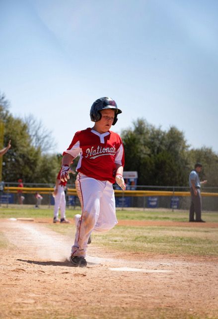 Young baseball player in red uniform running along baseline during youth baseball game on sunny day.