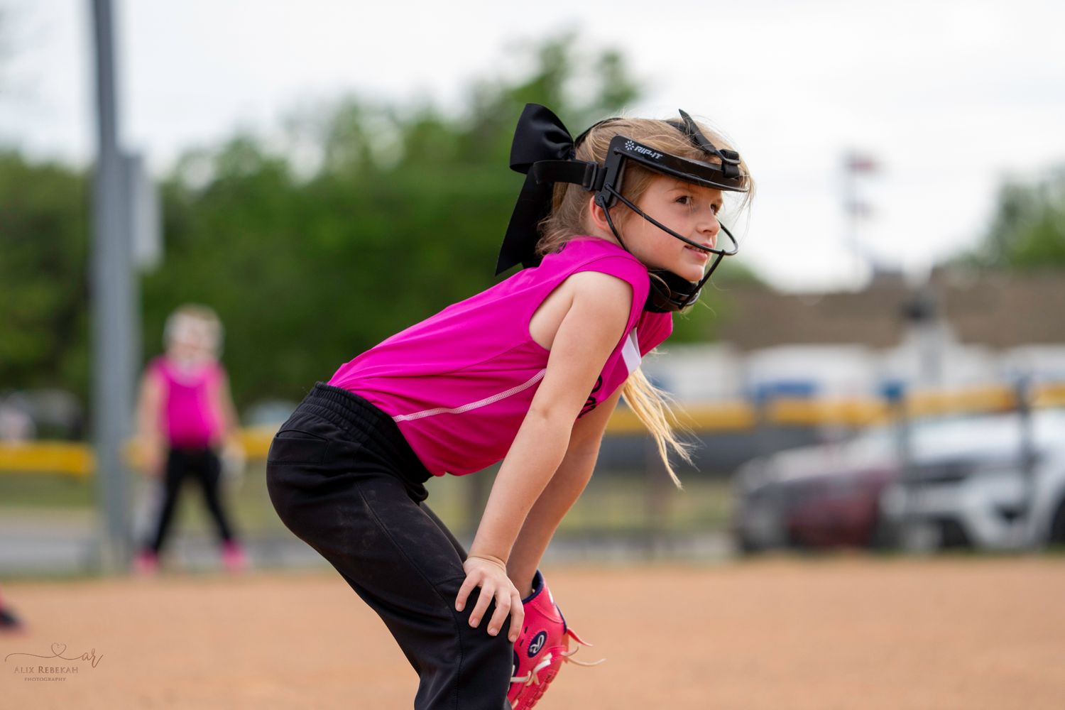 Youth softball player in pink jersey and black pants stands ready in fielding position on dirt infield.