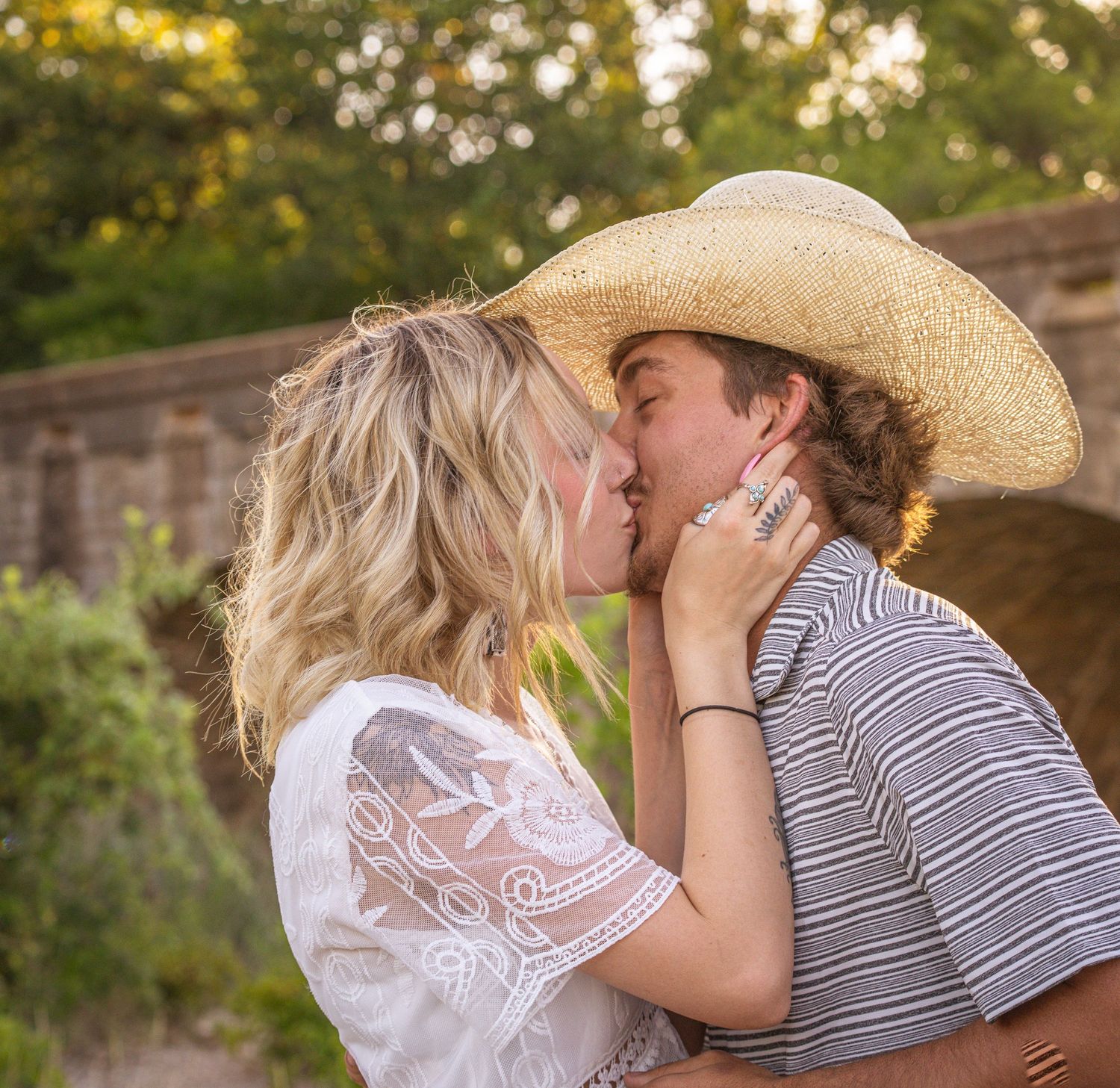Romantic couple shares tender kisses in soft outdoor lighting, with a woman in white lace and man in striped shirt.
