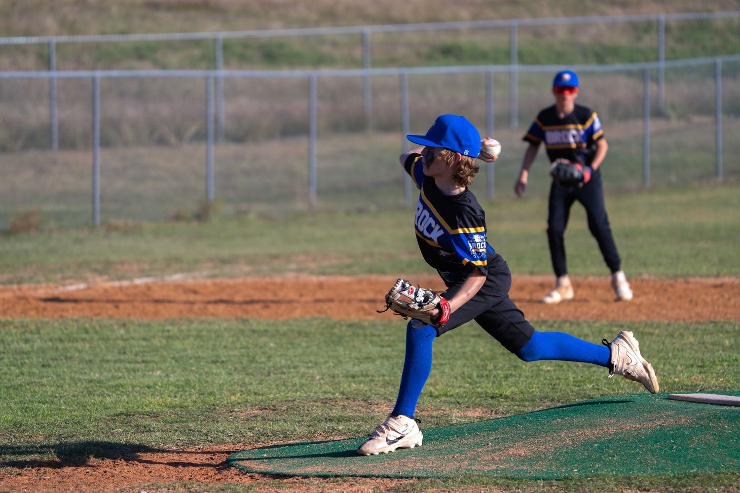 Youth softball pitcher in blue uniform winds up for a pitch on dirt mound during game practice.
