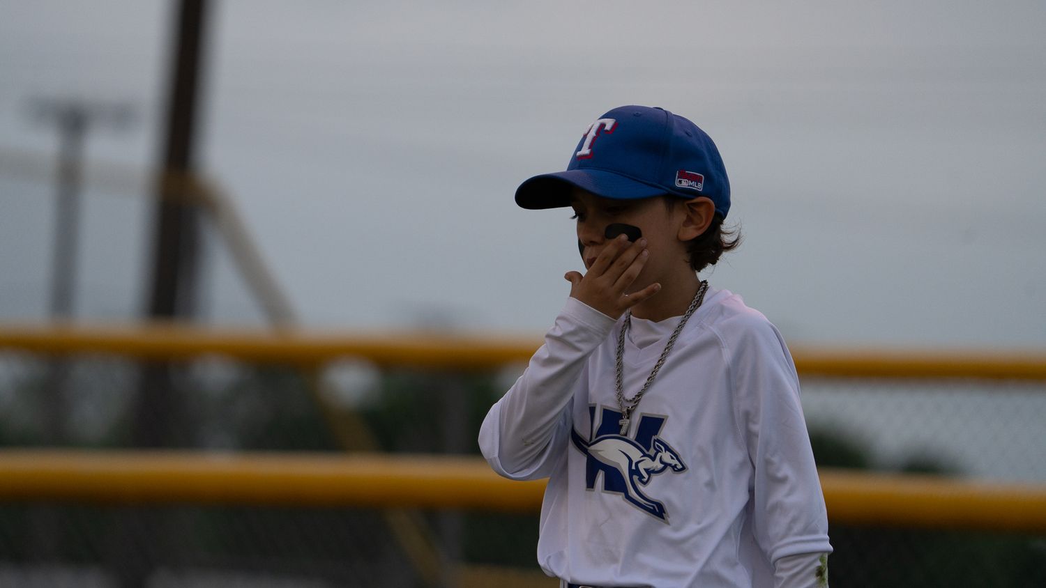 A baseball player wearing a white uniform with blue cap and collar stands on a ballfield in overcast weather.