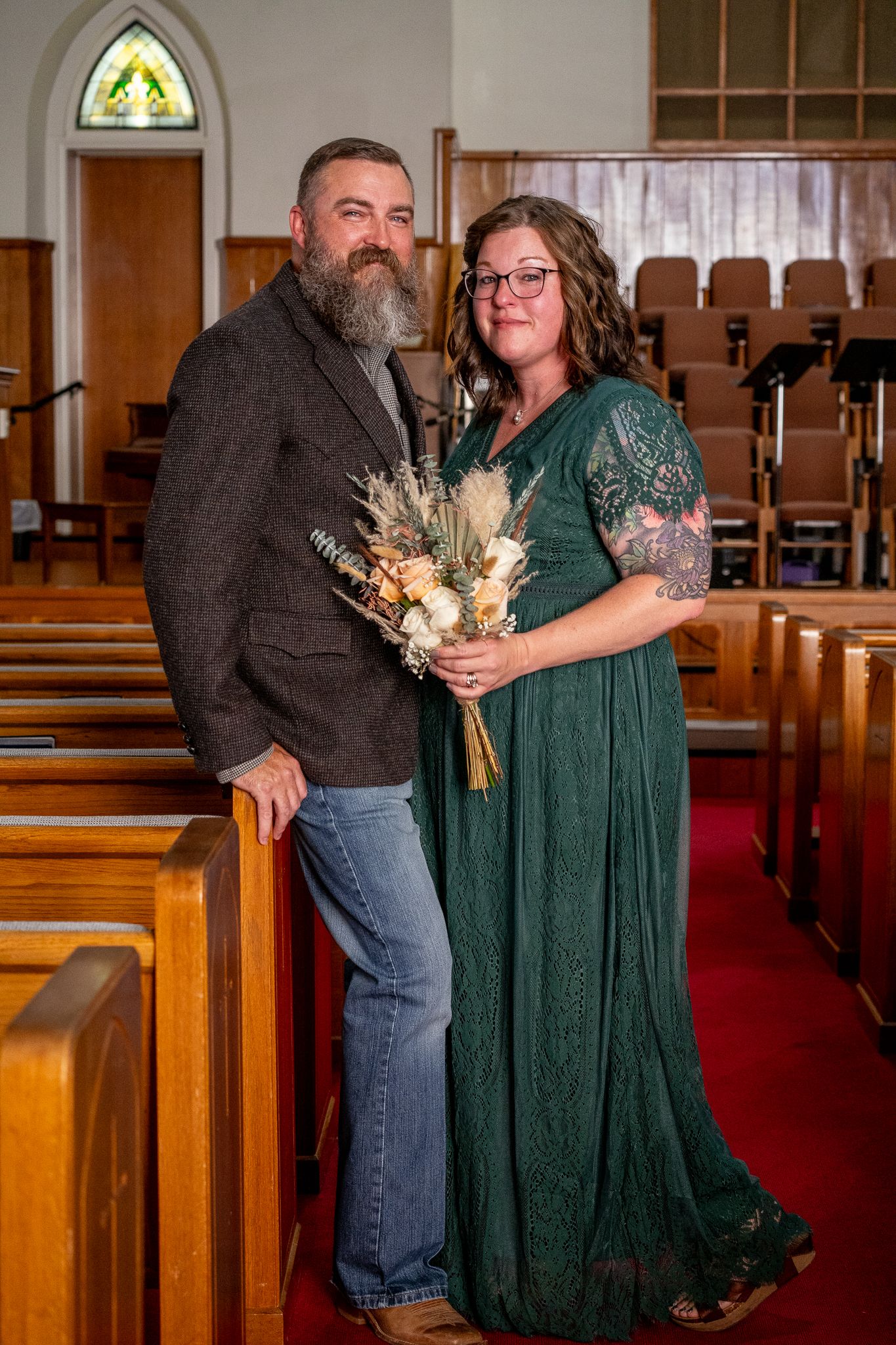 Wedding portrait in church with wooden pews and stained glass window.