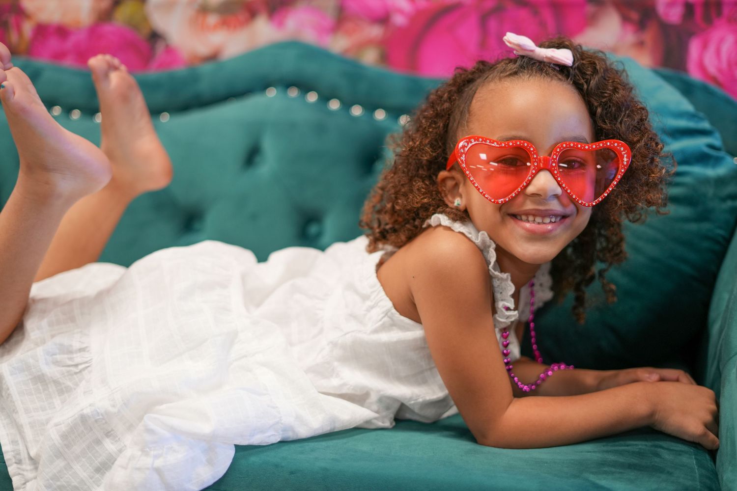 White dress and pink heart glasses complement a relaxed pose on emerald velvet couch.