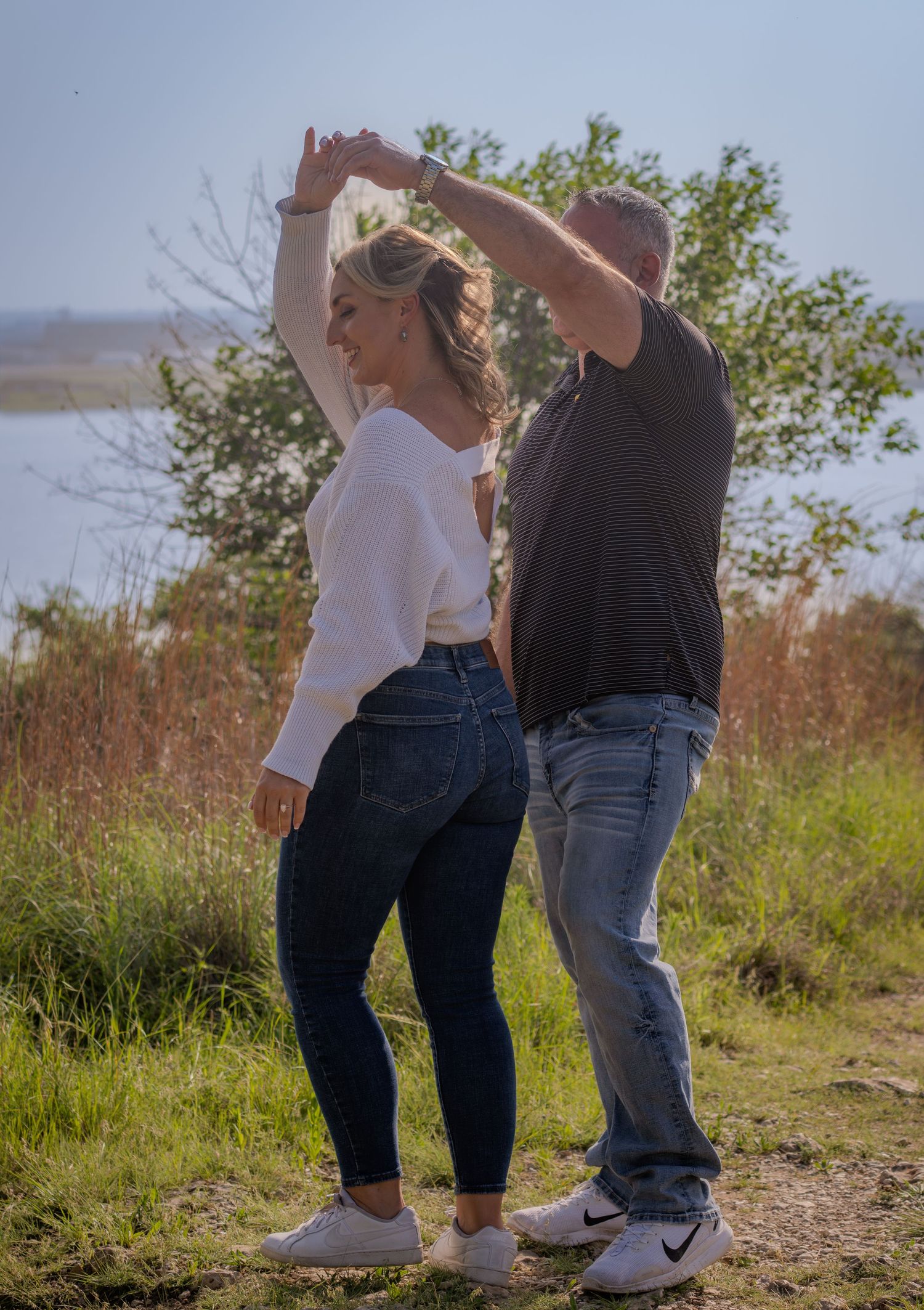 Couple dancing outdoors in jeans, woman in white top being twirled by man in black shirt near a scenic lake.