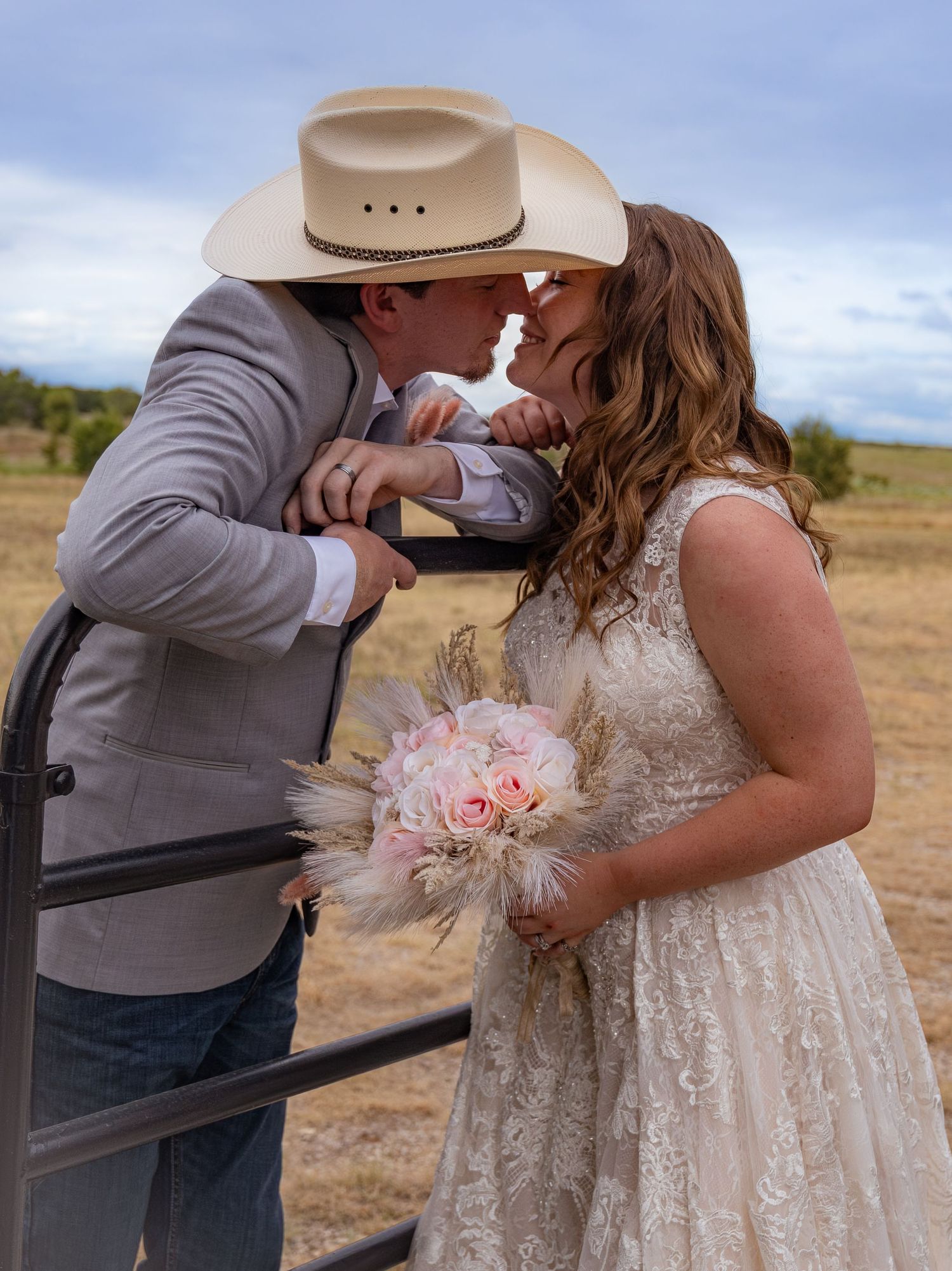 A couple shares a kiss at their rustic outdoor wedding, with the groom in a cowboy hat and bride holding a pink bouquet.
