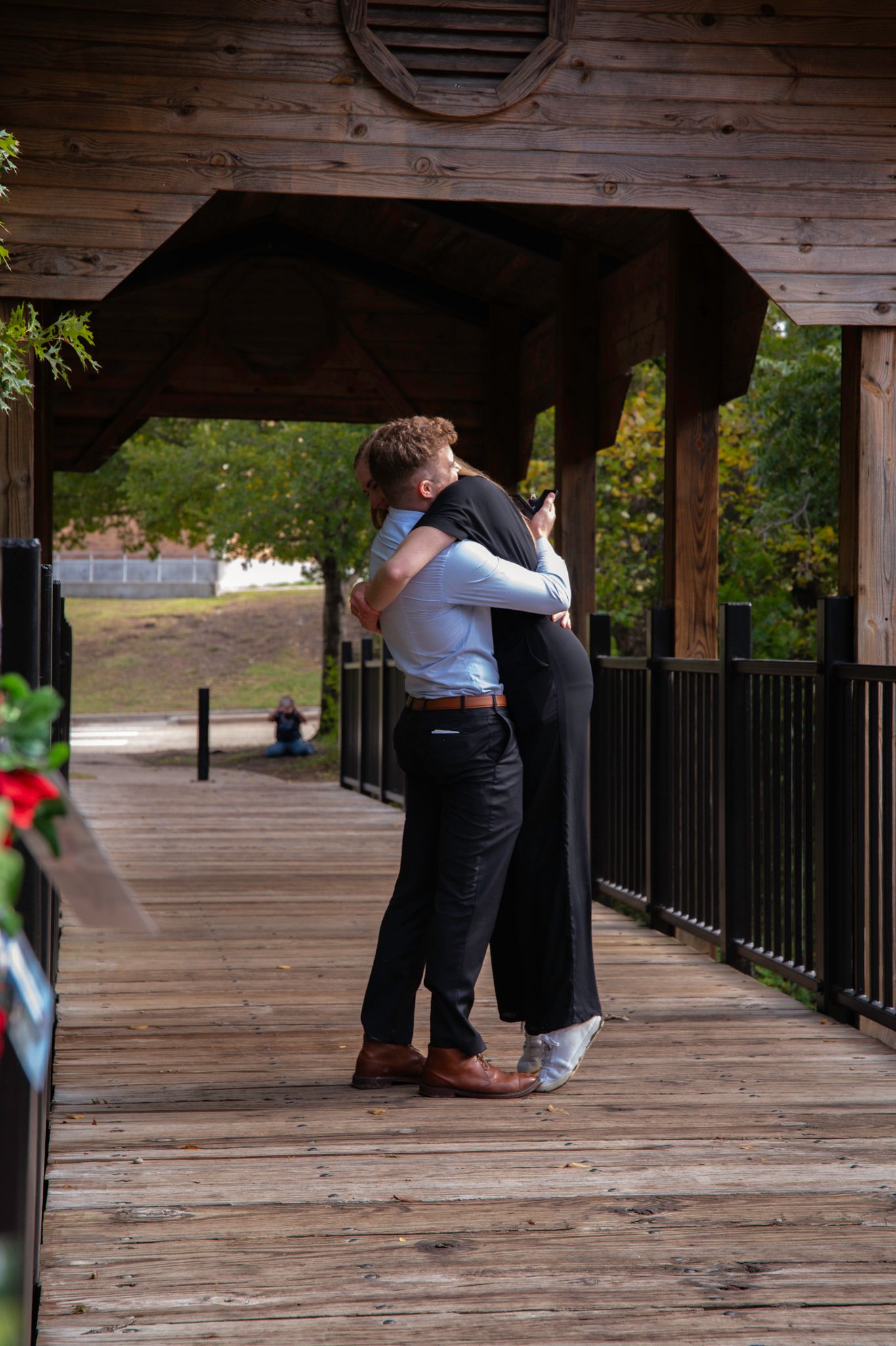 Two people share a tender embrace on a wooden covered bridge surrounded by greenery.
