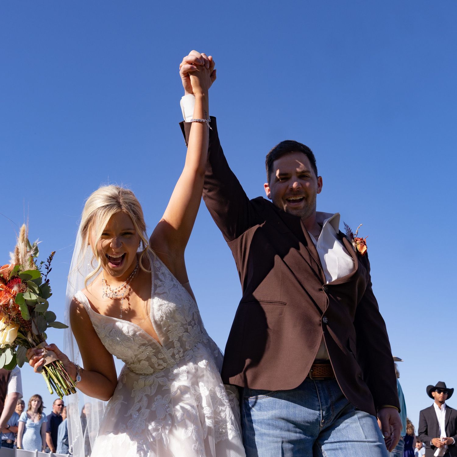 A couple celebrating their wedding moment outdoors with raised arms against a bright blue sky.