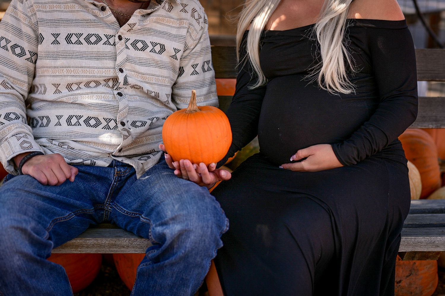 Two people sit on a bench with a pumpkin during an autumn photo session.