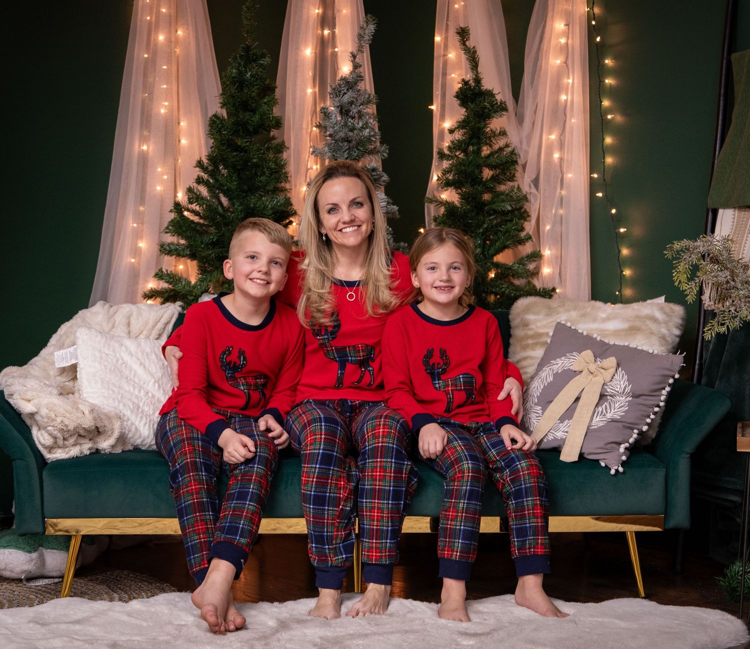 Matching family in red reindeer sweaters and plaid pajama pants sitting on green couch with Christmas trees and lights.