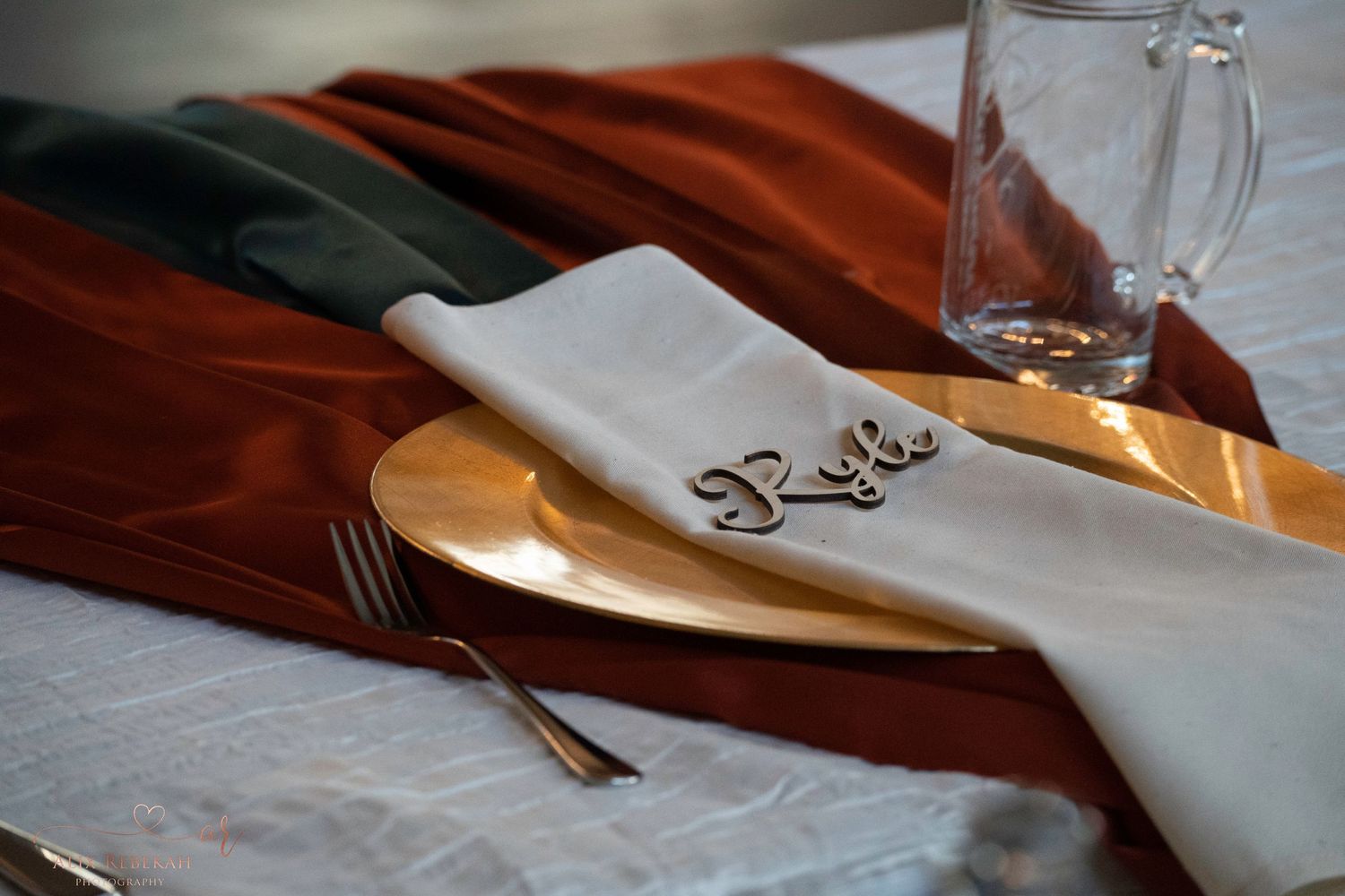 Elegant place setting with gold charger plate and white napkin on burgundy tablecloth.