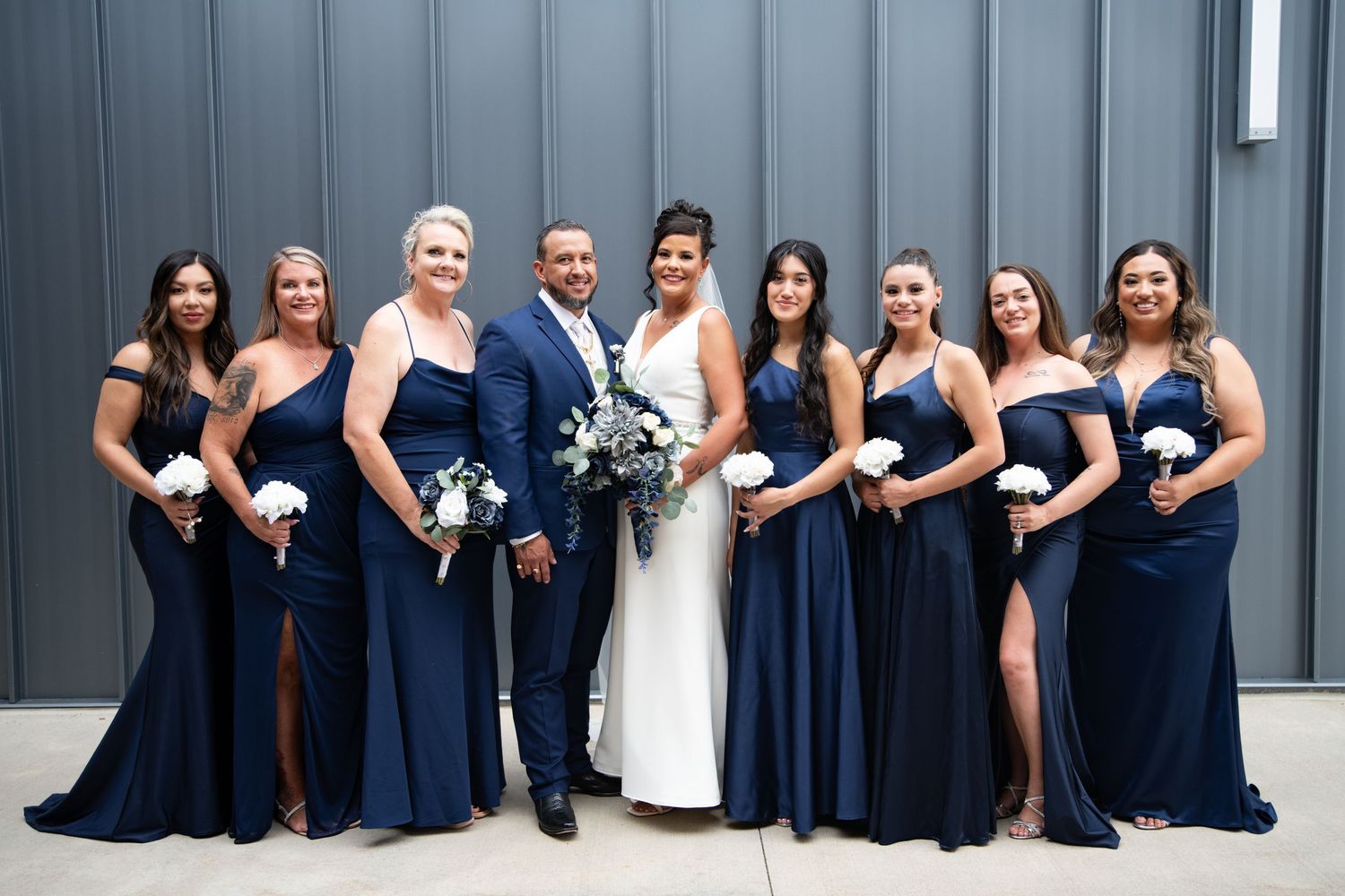 Wedding party in navy blue gowns and grey suits poses against industrial grey wall.