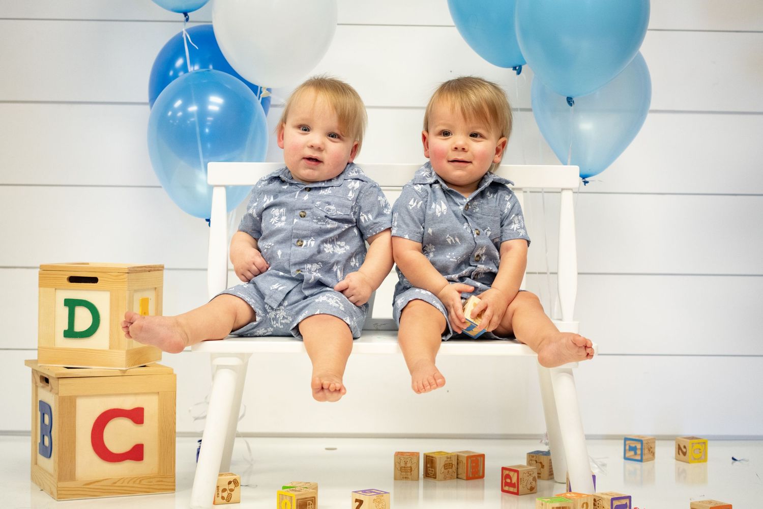 Twin babies in gray rompers sitting on white bench with letter blocks and balloons.