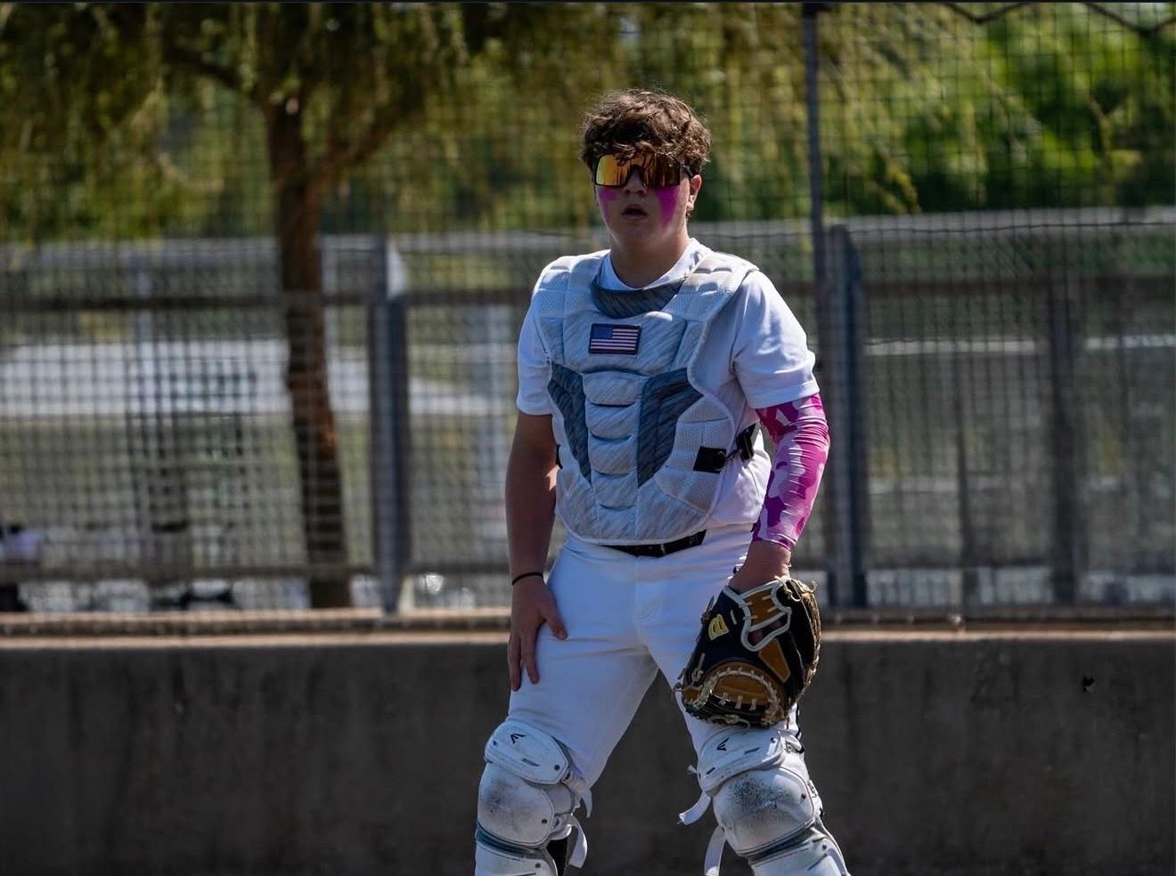 Baseball catcher in white uniform and chest protector holding a mitt on an outdoor field.