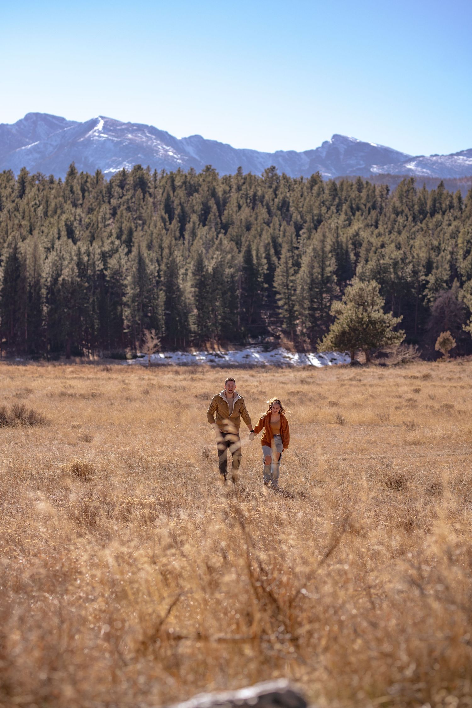 Two people walking through a golden meadow with snow-capped mountains and pine forests in the background.