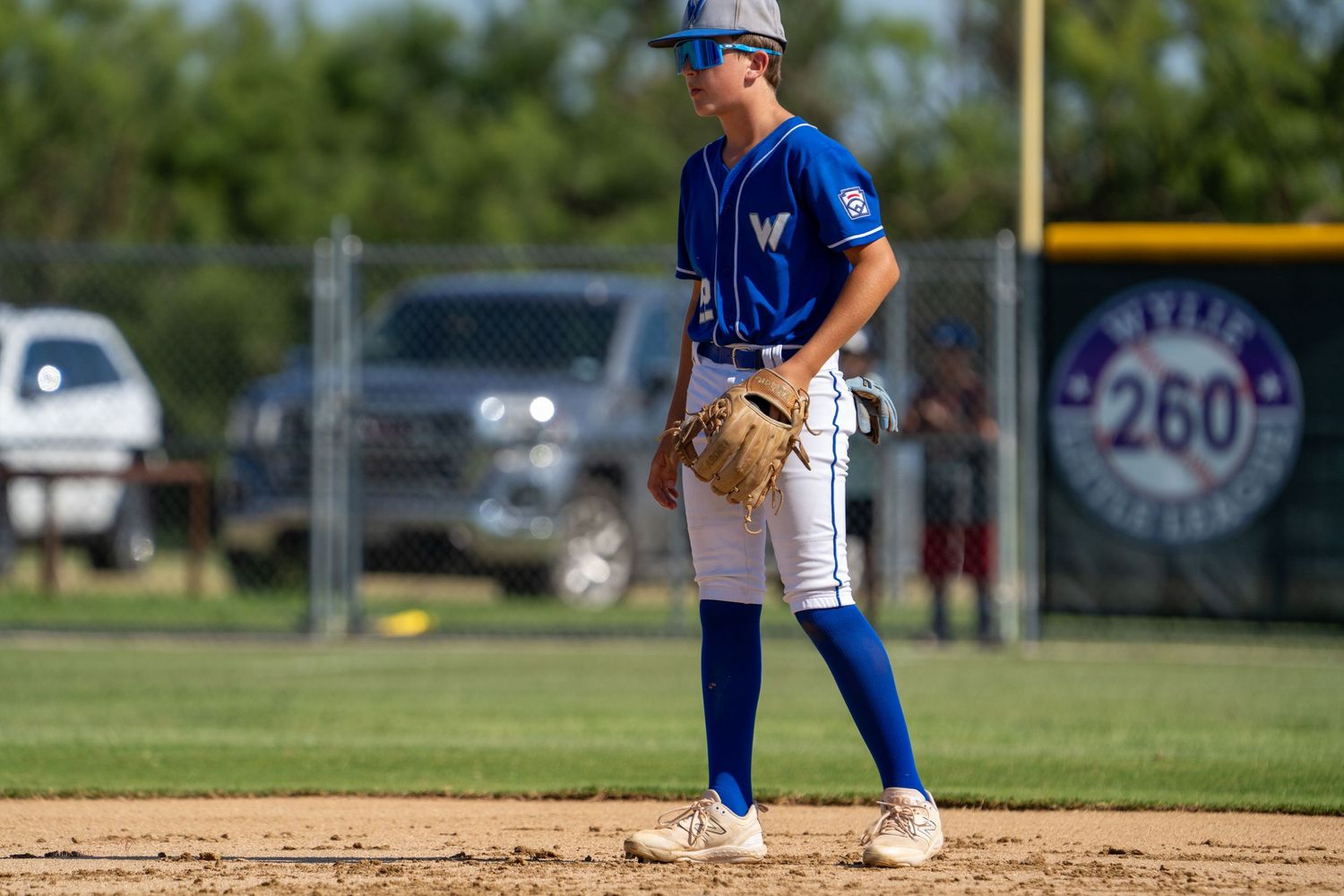 Baseball player in blue uniform and cap stands ready on the base during a game with field and fence visible in background.