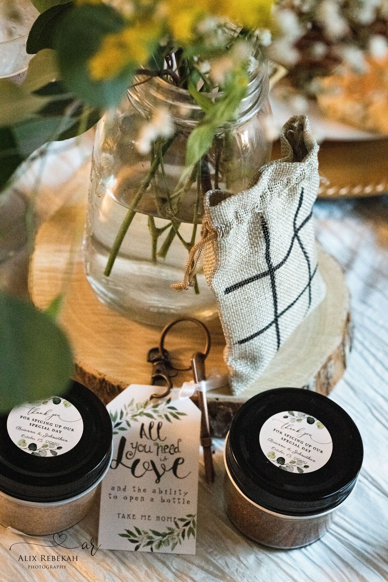 Mason jars with wedding favors and rustic decor display natural body scrubs with handwritten gift tags on wooden surface.