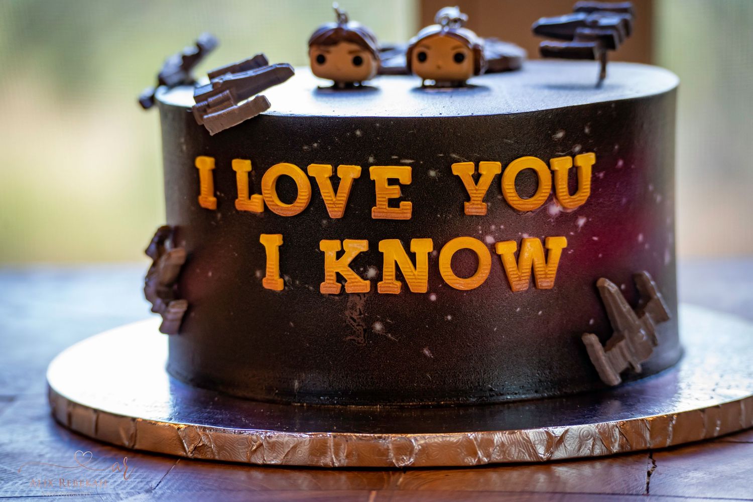 A dark chocolate wedding cake with gold text reading 'I LOVE YOU I KNOW' on a wooden board.