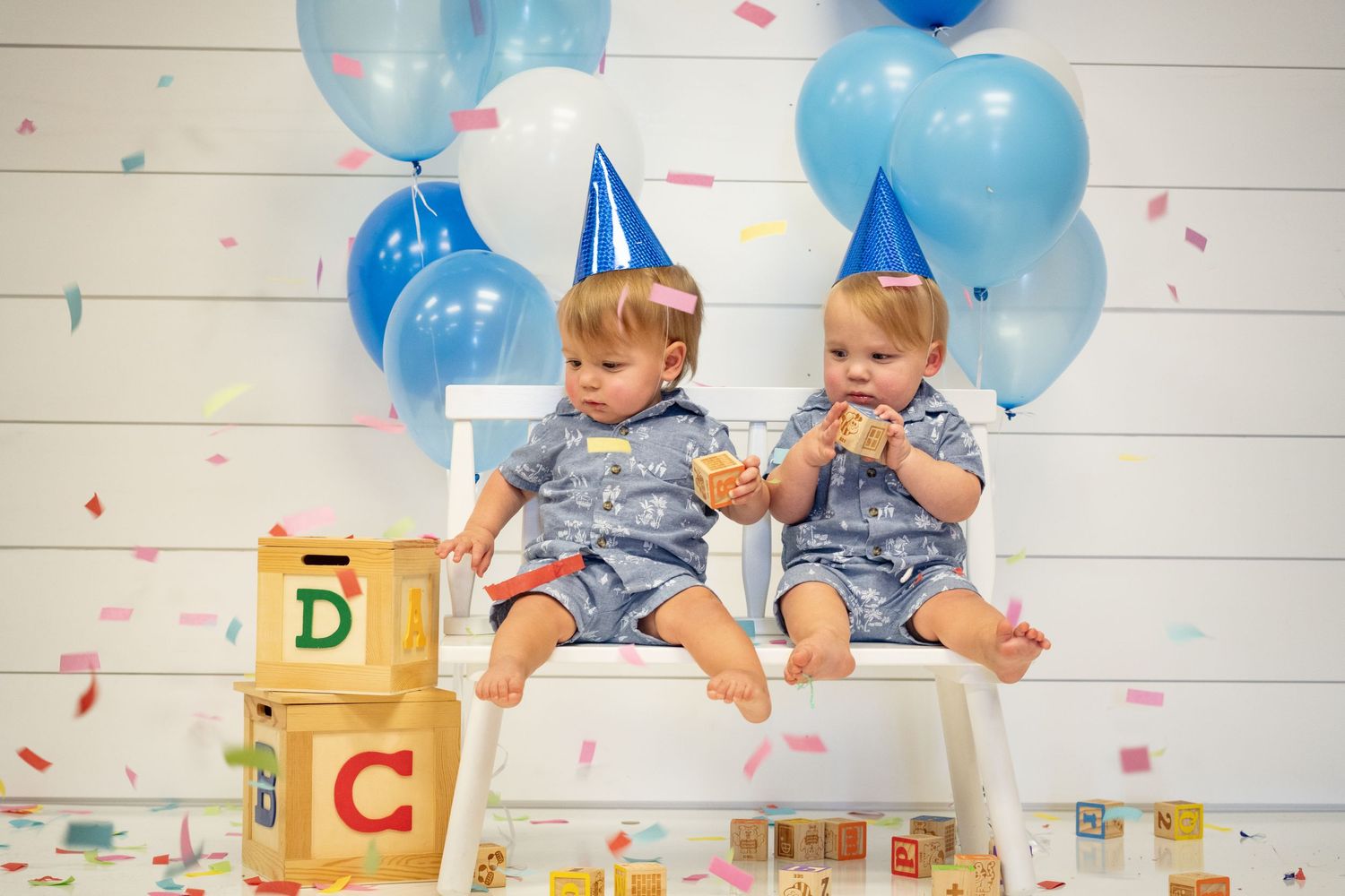 Twin babies celebrate with party hats, blue balloons and confetti.