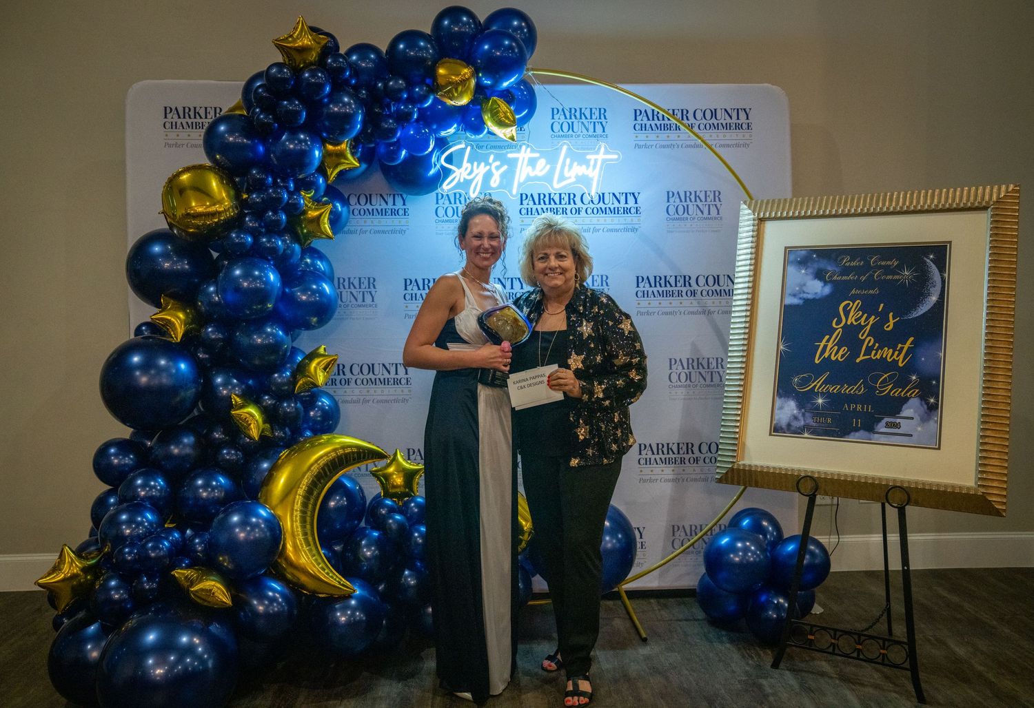 Two people pose in front of a balloon arch with blue and gold decorations at an awards ceremony.