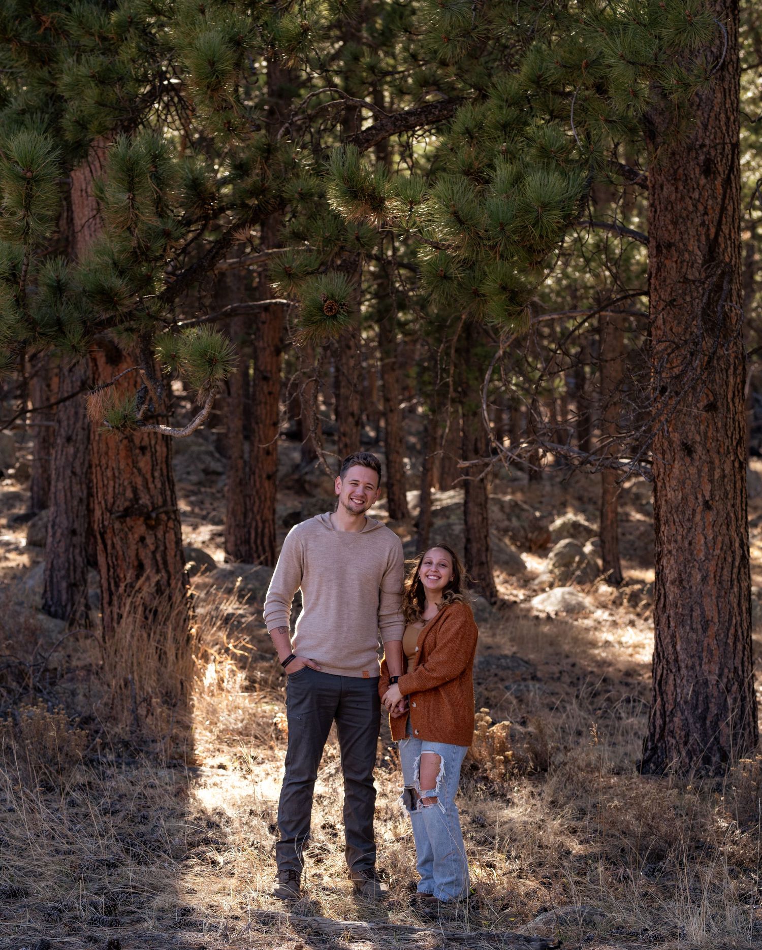 A couple stands together among tall pine trees in a forest setting wearing casual autumn attire.