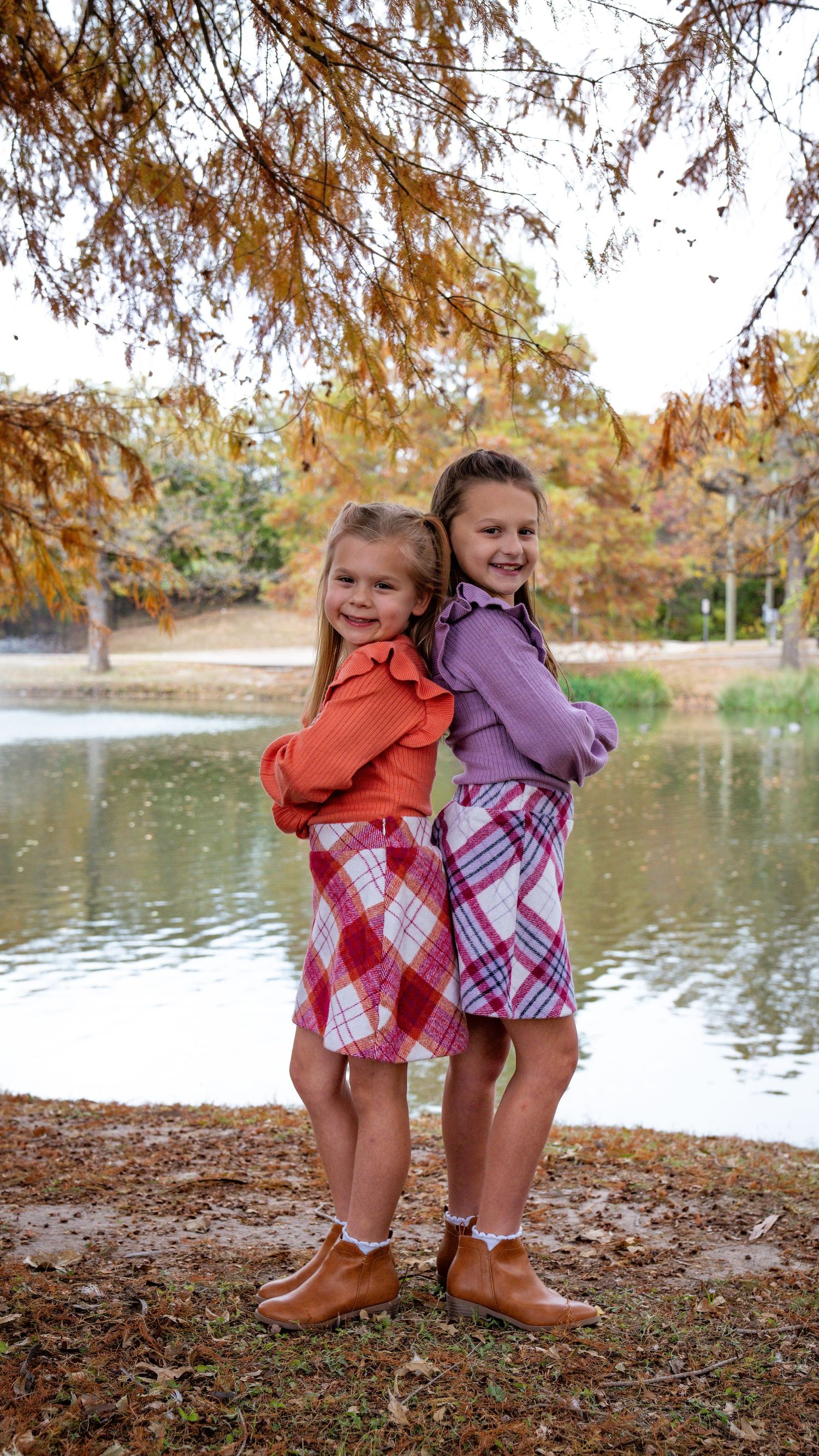 Two young children in plaid skirts and jackets pose back-to-back near a lake during autumn with fall foliage in background.