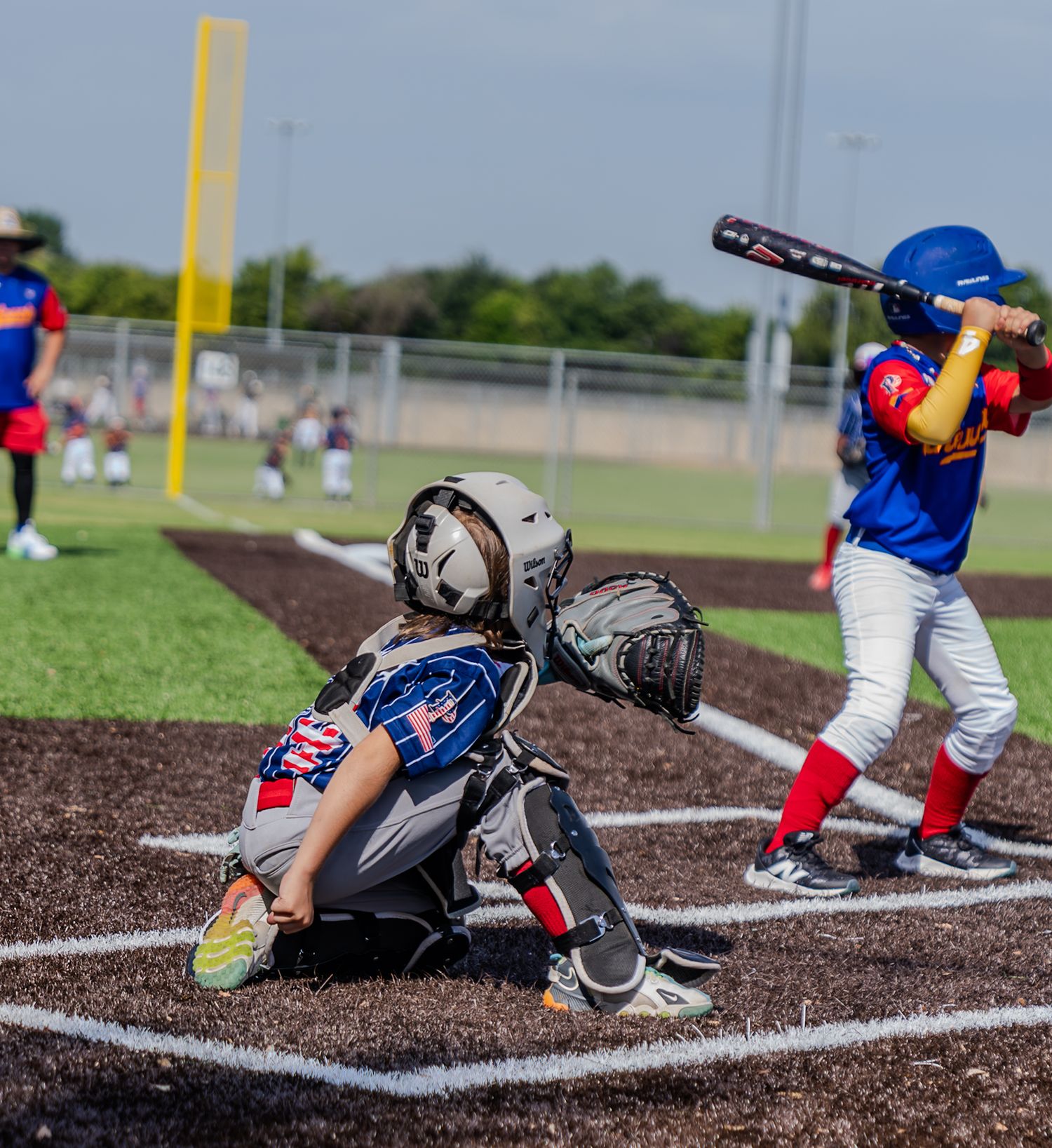 Young baseball players in action, catcher crouching at home plate while batter prepares to swing during youth league game.
