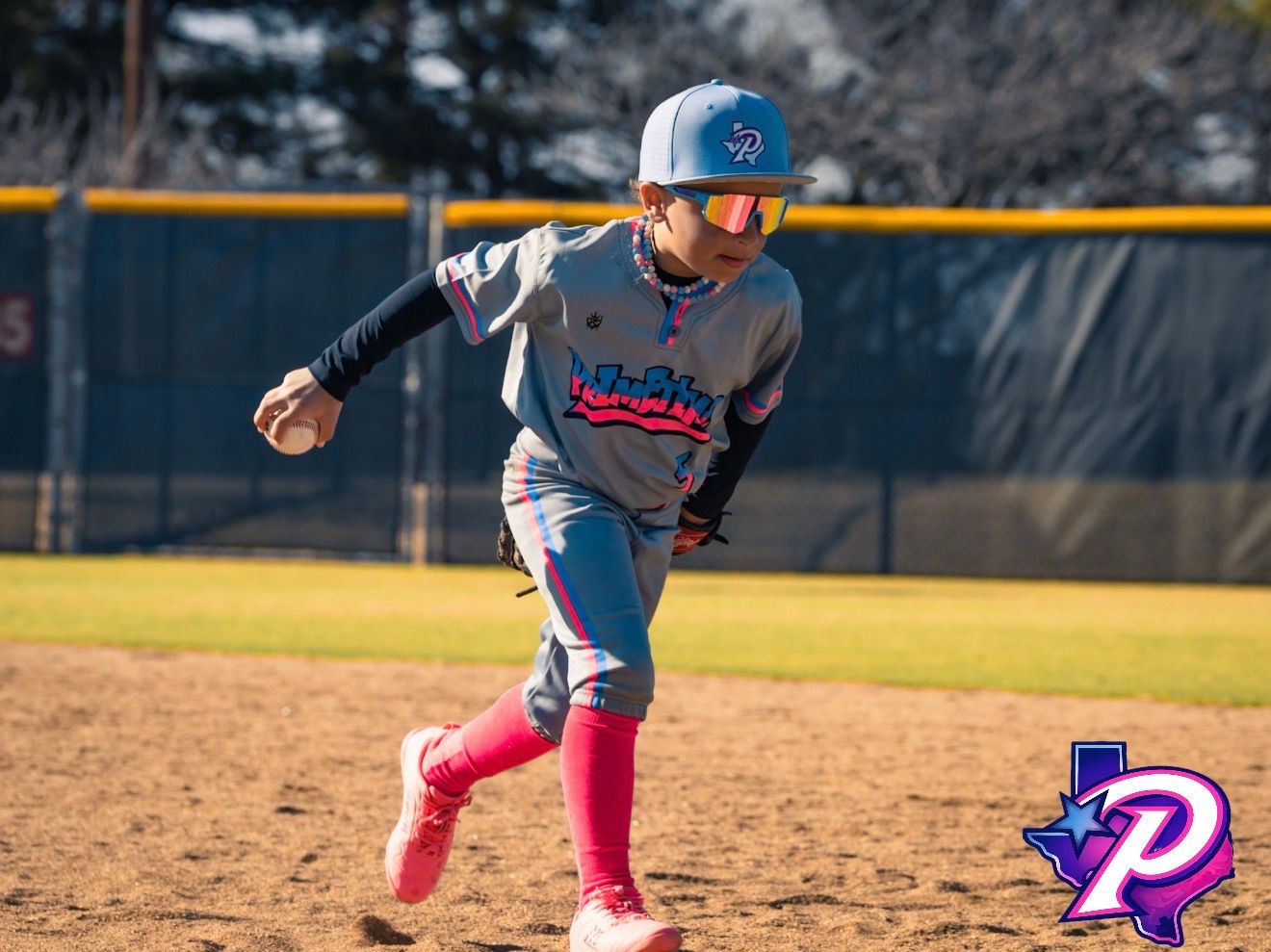 Young baseball player in gray and pink uniform running bases during a sunny outdoor game.