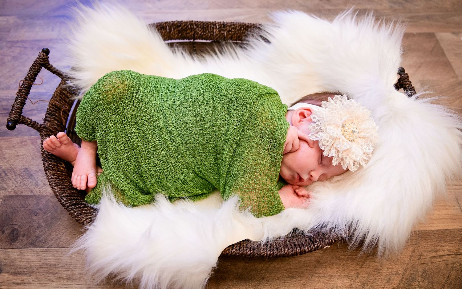 Sleeping baby wrapped in green knit on white fur in rustic basket.