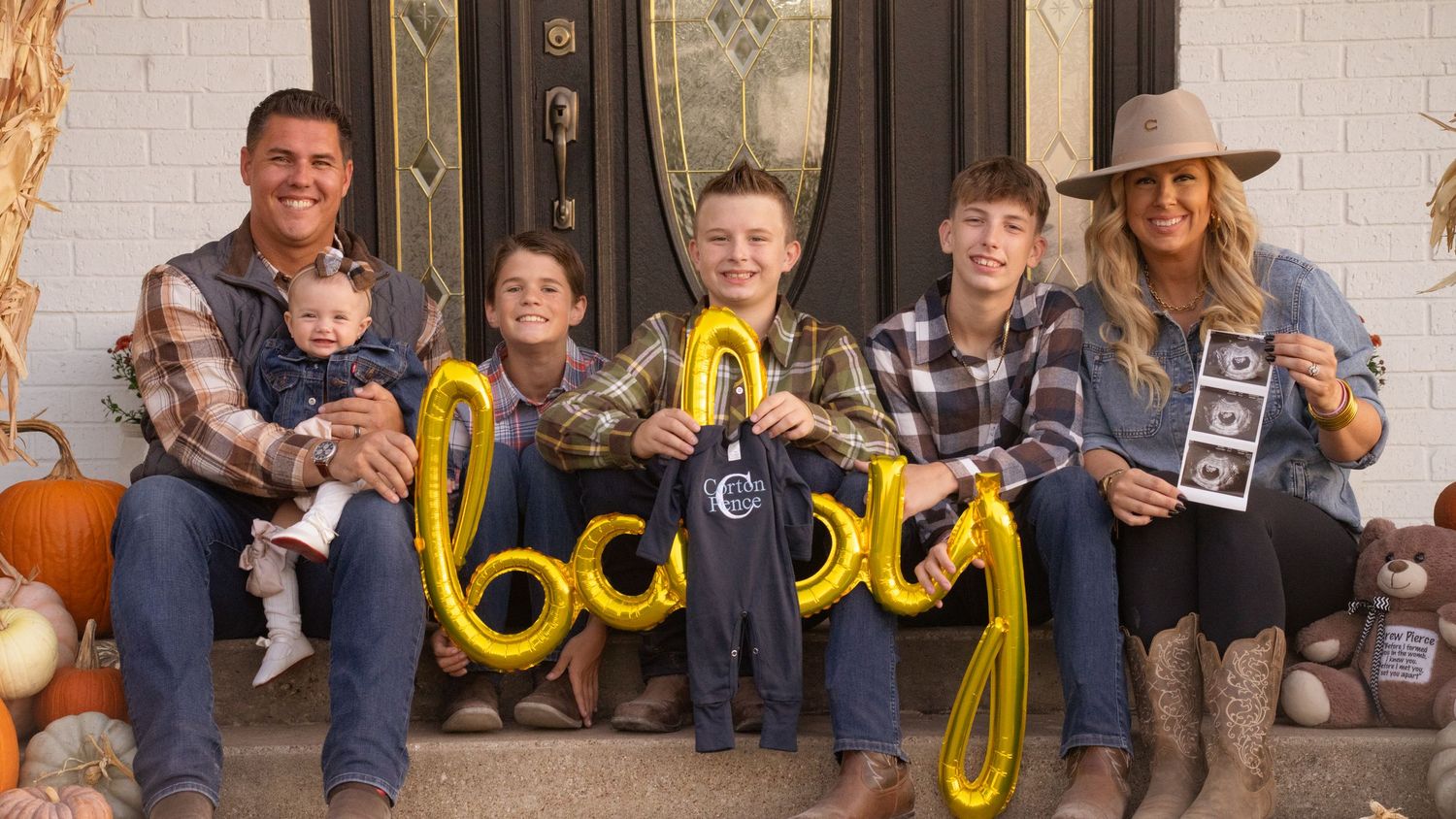 Group sitting on porch steps holding gold balloon letters spelling 'baby' during fall announcement photo.
