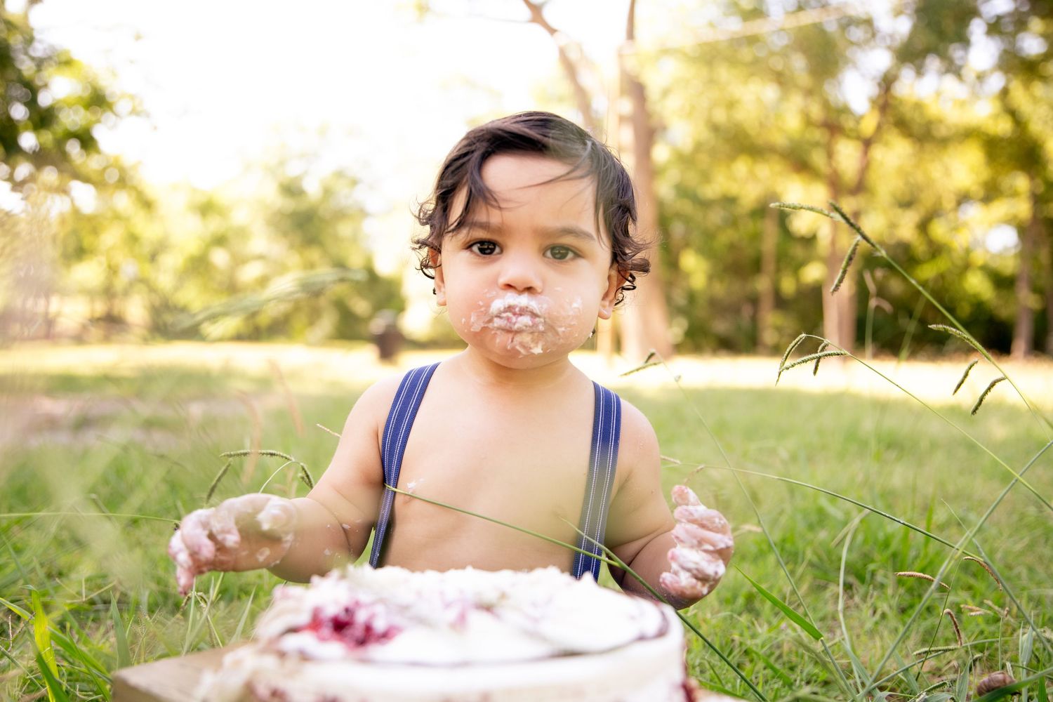 A small child in blue overalls playing with cake outdoors during a smash cake photoshoot.