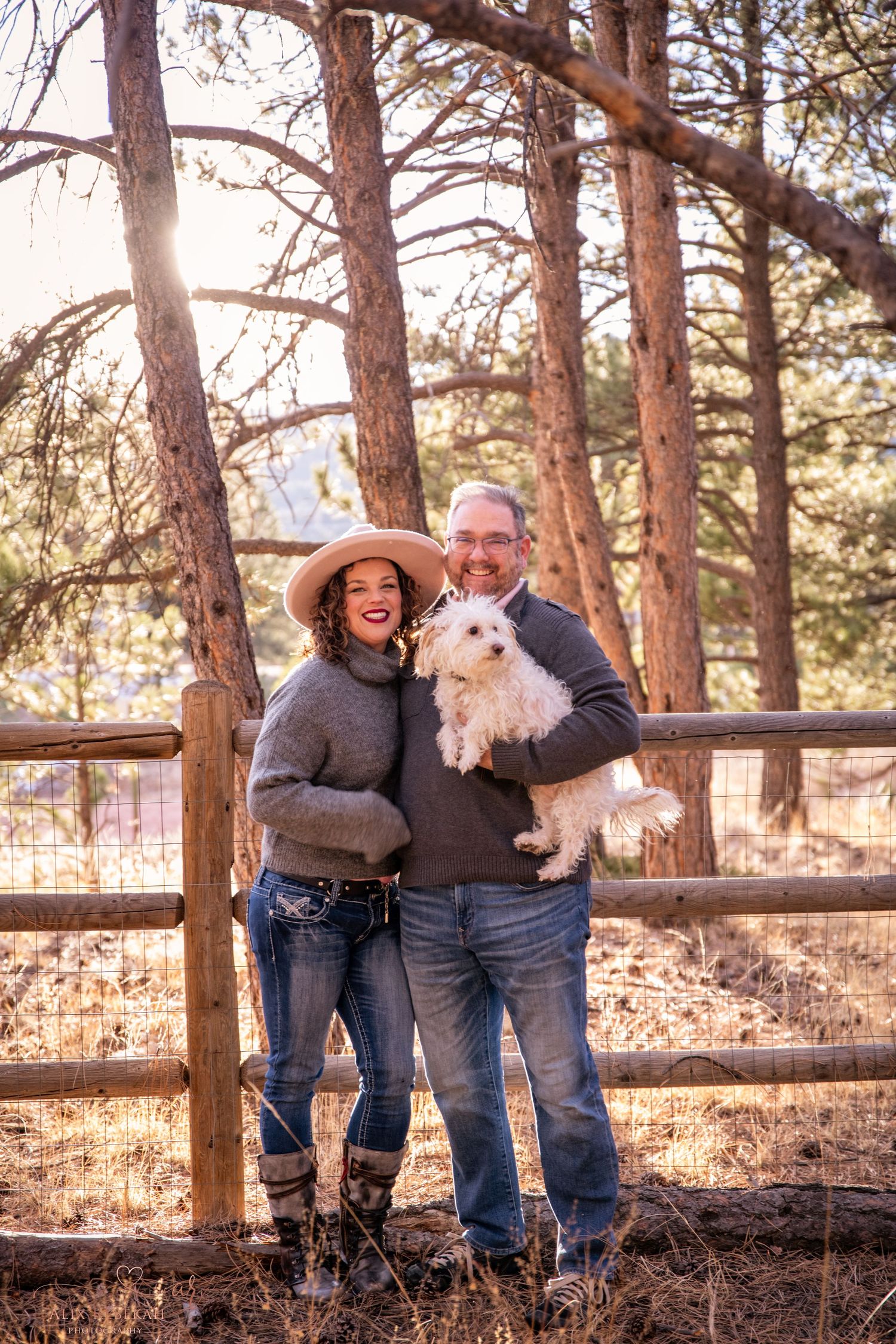 A couple stands with their small white dog against a wooden fence in a pine forest.