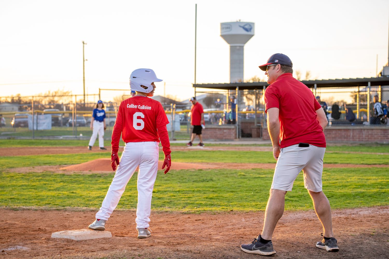 Youth baseball player stands on base while coach gives instruction during practice on sunny evening at baseball field.