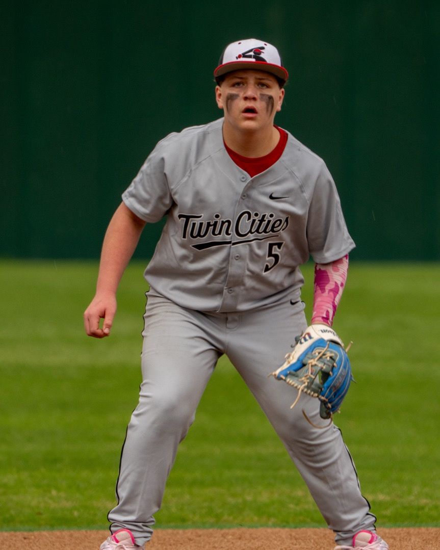 Twin Cities baseball player #5 in gray uniform crouches in ready position on field wearing pink batting gloves.