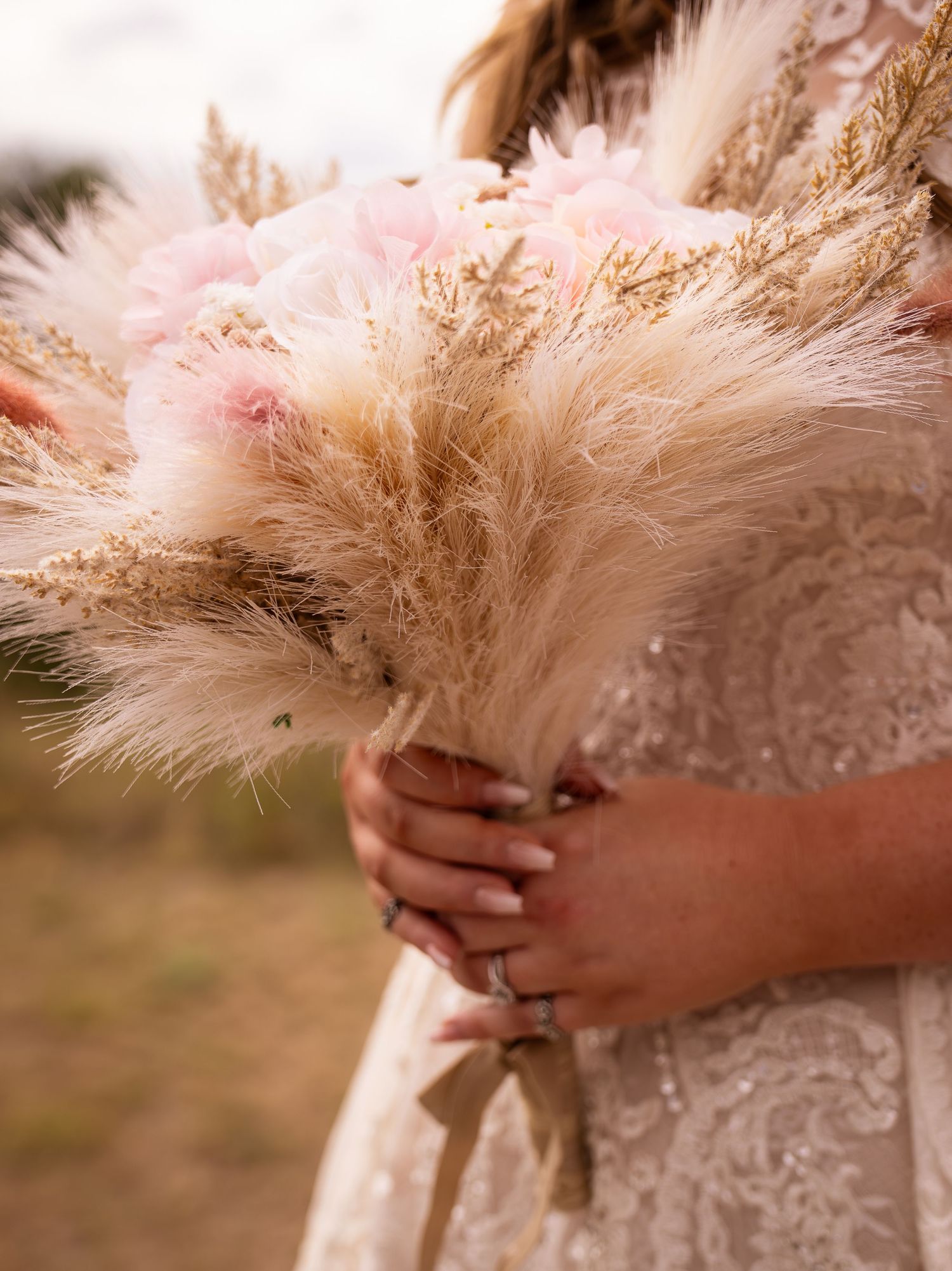 Bride holding a boho wedding bouquet of pampas grass and pink flowers, wearing a lace gown with a ring on her finger.