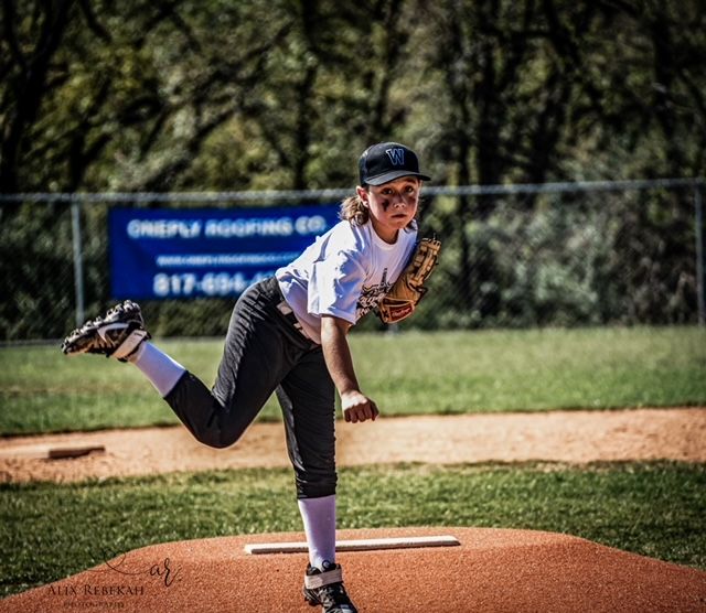 Youth baseball pitcher in follow-through motion on dirt mound during game with trees in background.