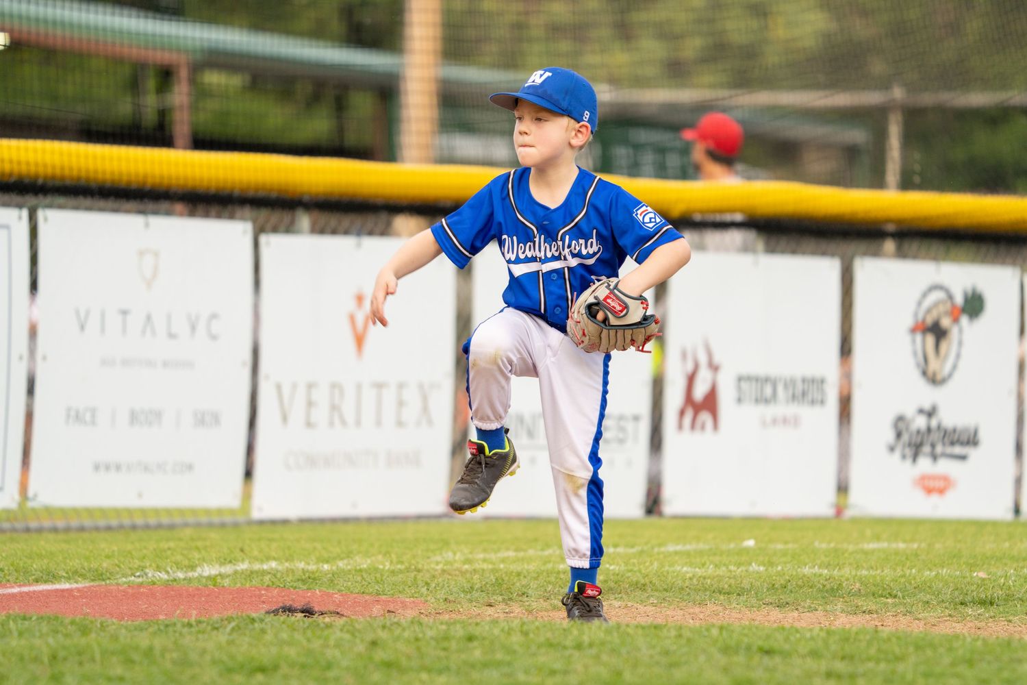 Baseball player throwing a pitch on a field with advertising banners visible along the outfield fence.