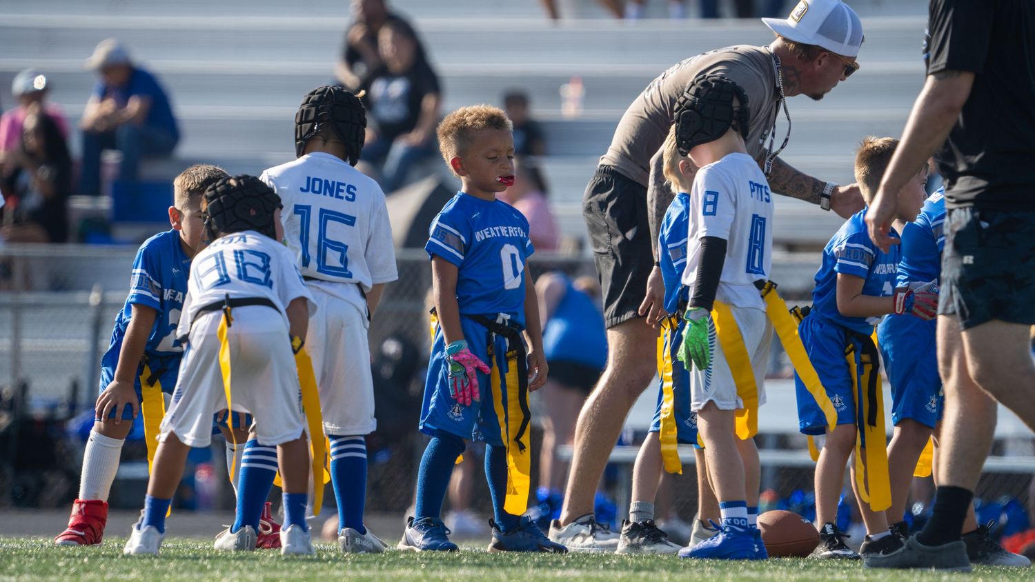 Youth football players and coaches huddle on the sidelines during a game wearing blue and yellow uniforms.
