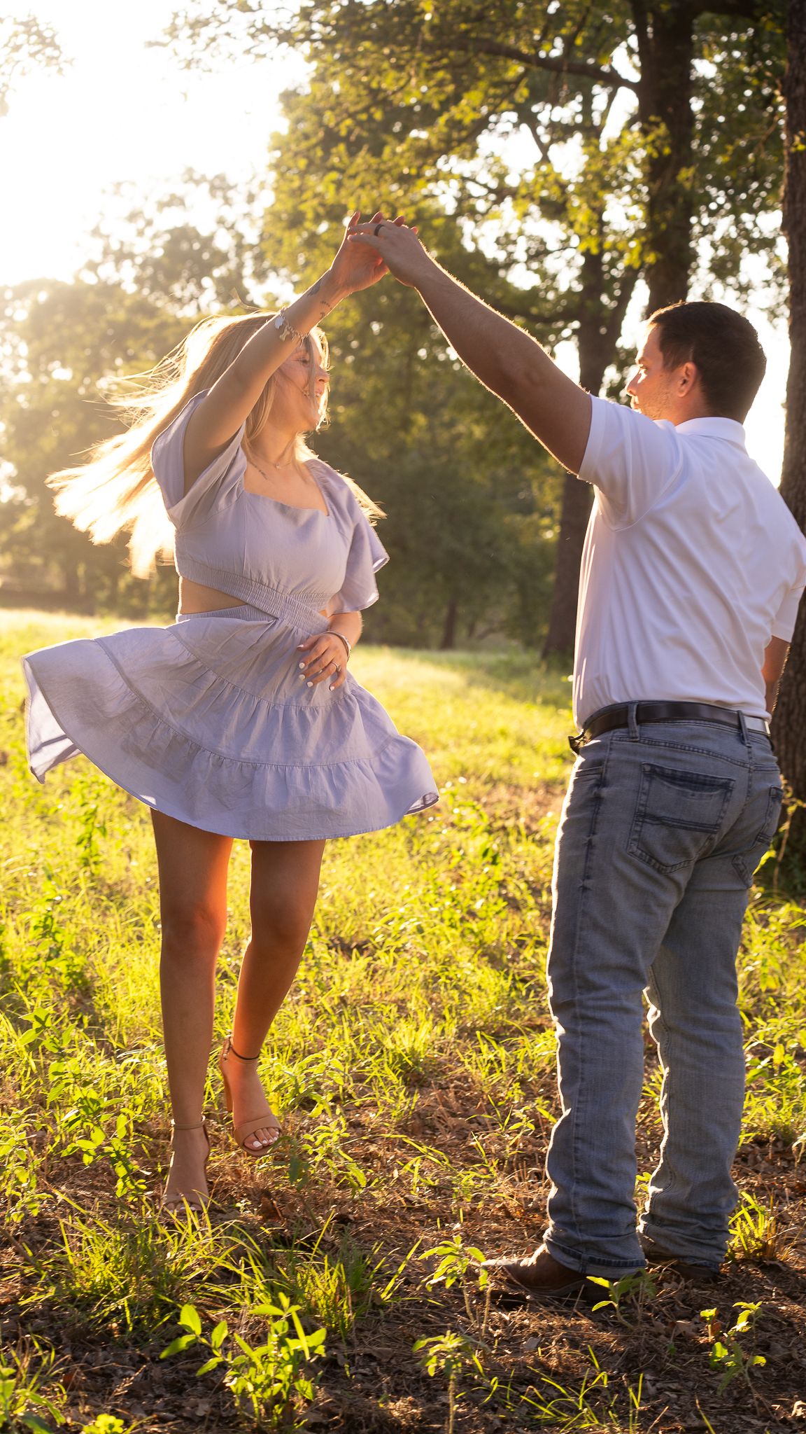 Couple dancing together in a sunlit meadow during golden hour while wearing white dress and casual attire.