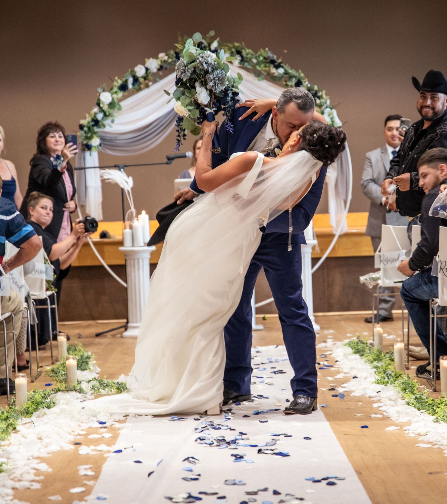 A romantic dip kiss moment during a wedding ceremony under a floral arch with scattered petals on the aisle.