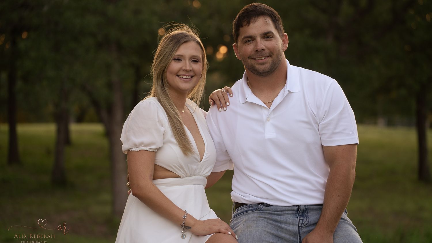 A couple in coordinating white outfits poses together in a natural outdoor setting.