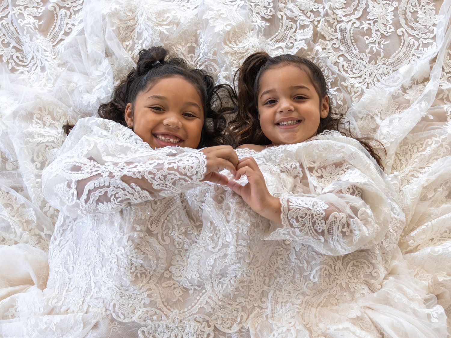 Two smiling girls lying on white lace fabric, making a heart shape with their hands together.