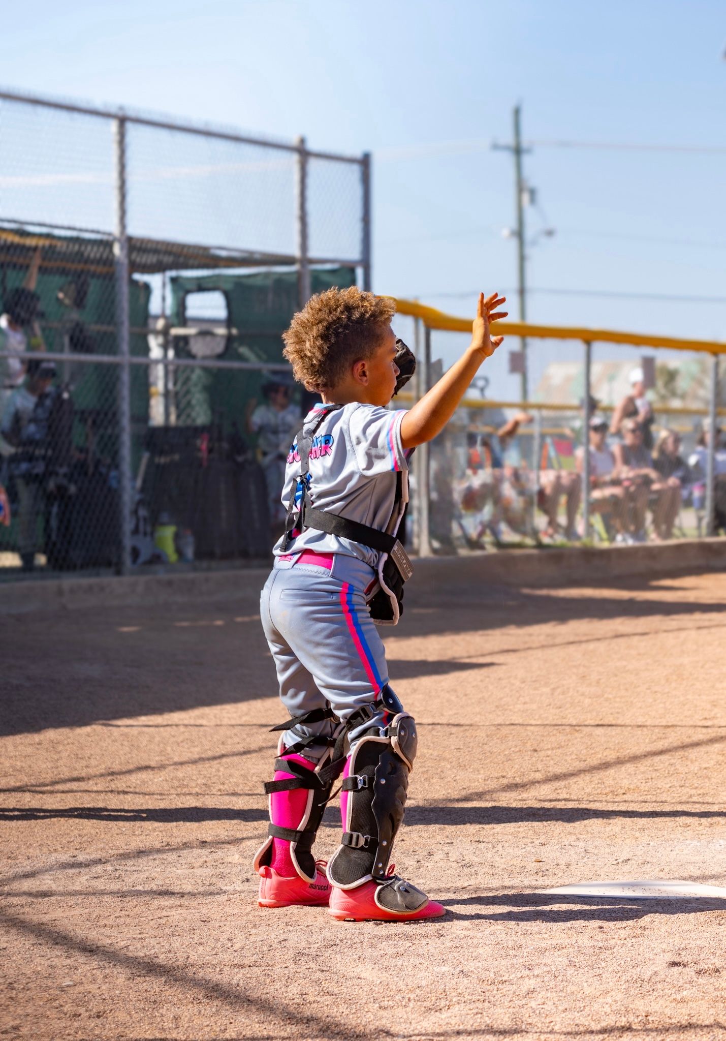 Young baseball catcher in pink and gray uniform with gear, reaching up to catch a ball on a dirt field.