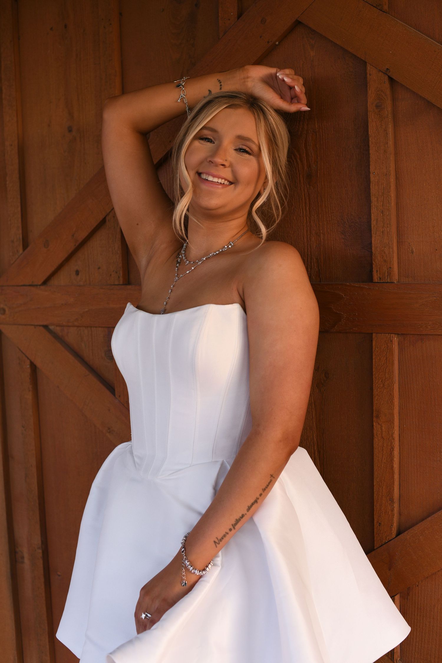 A joyful person wearing a white strapless dress poses against a rustic wood barn background with a warm smile.