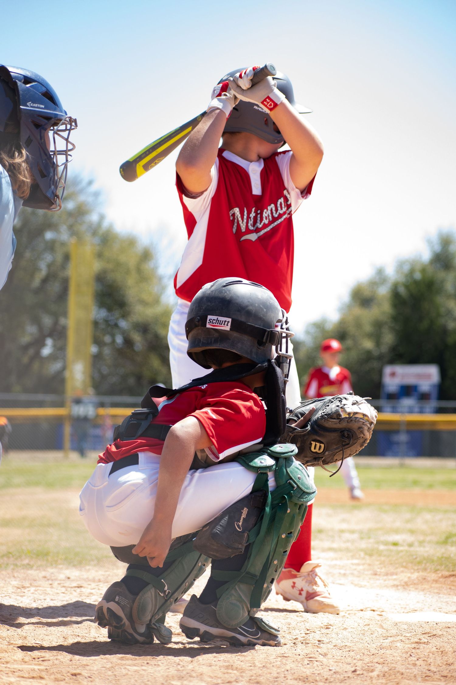 A baseball player in red uniform crouches behind home plate as a batter prepares for the pitch during a game.
