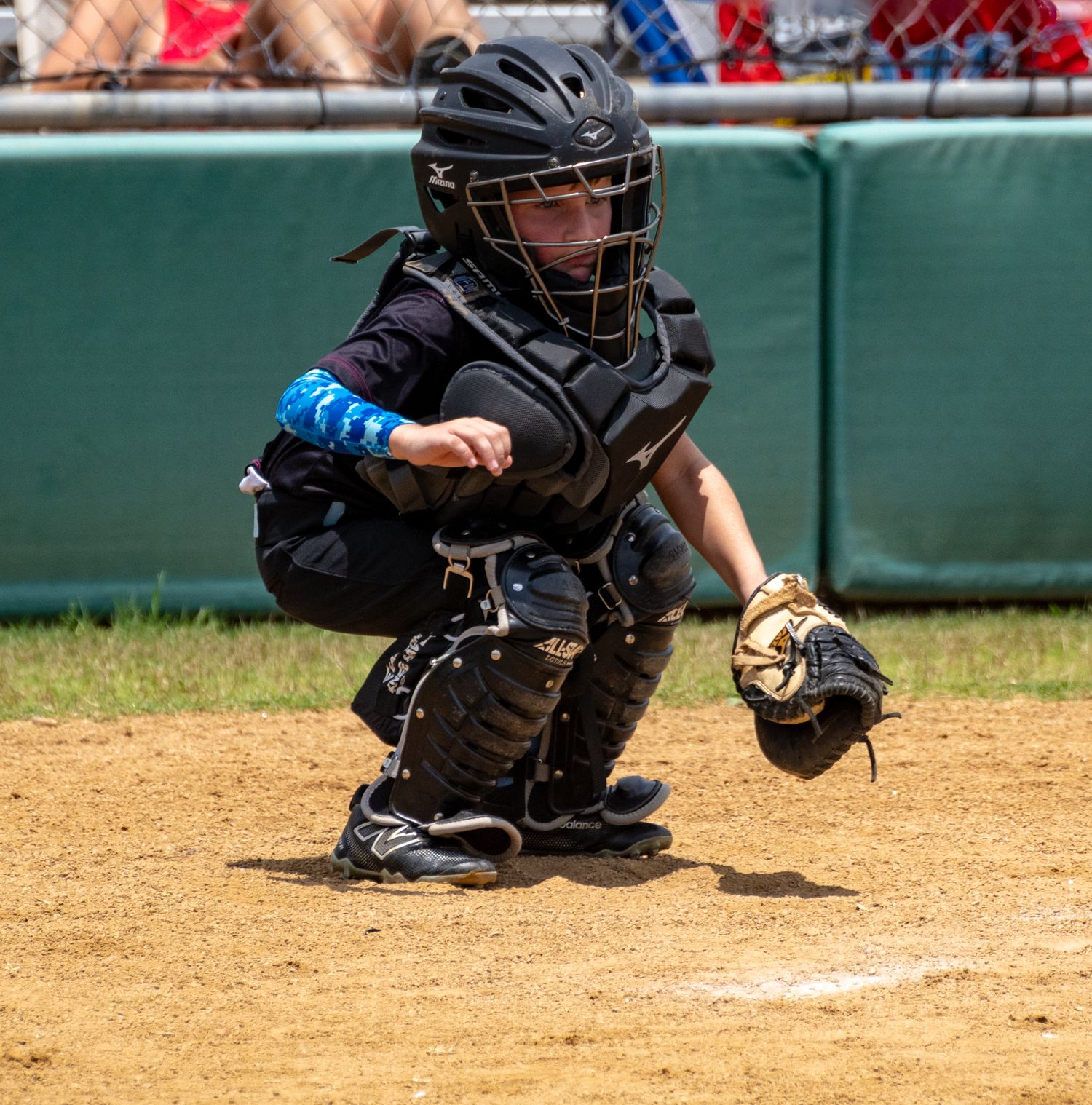 Young baseball catcher in black protective gear crouches in ready position behind home plate during youth game.