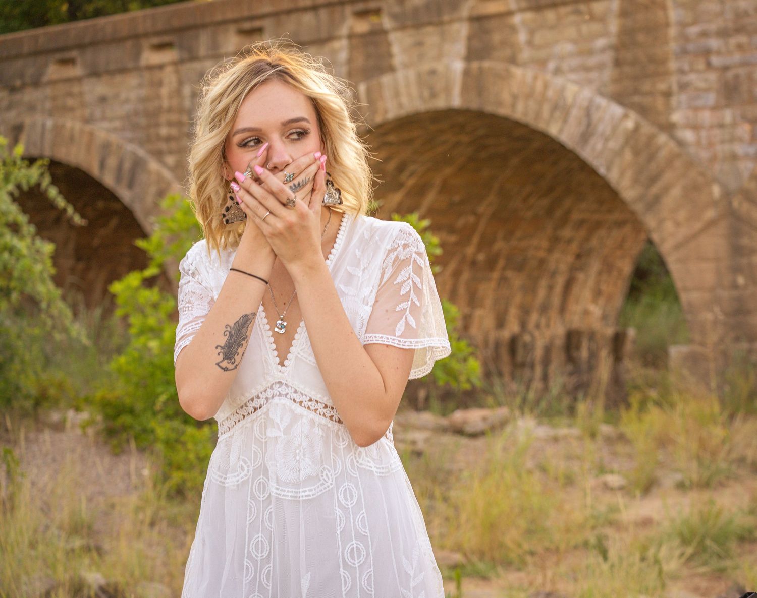 A woman in a white lace dress poses playfully with hands on face near a stone bridge in a natural setting.