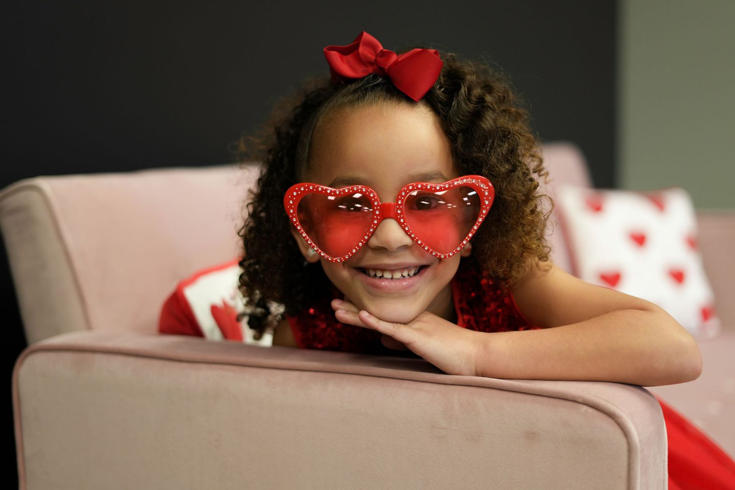 Curly-haired child in red dress and heart sunglasses poses playfully on a white couch.