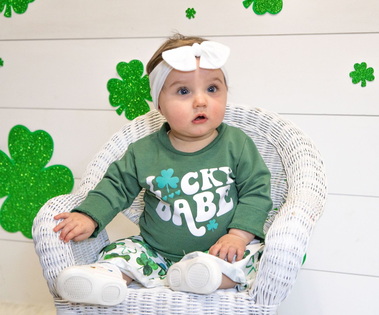 A baby wearing St. Patrick's Day themed clothing sits in a white wicker chair surrounded by green shamrock decorations.