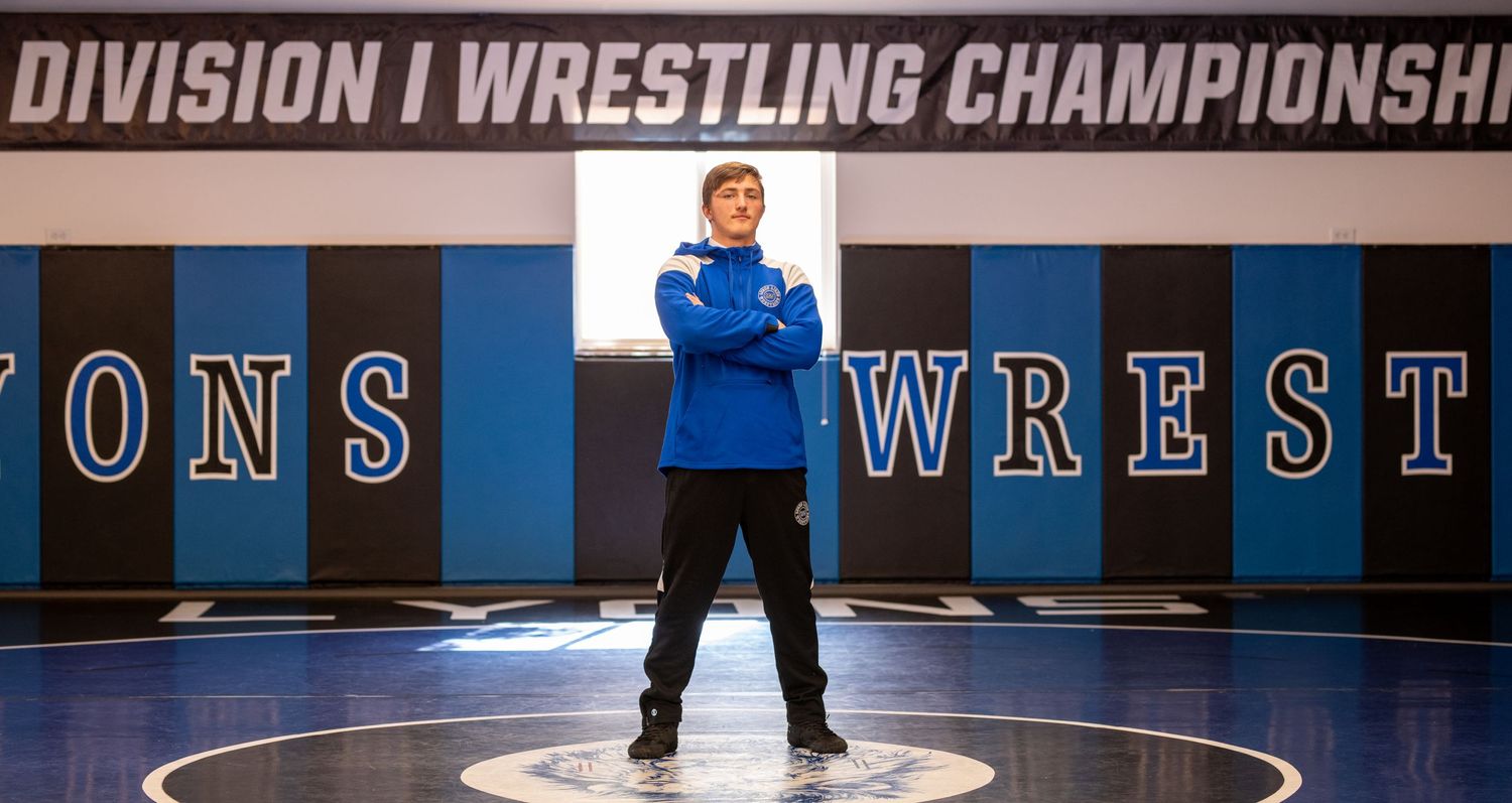 Athletic figure standing in center of wrestling mat under championship banner.