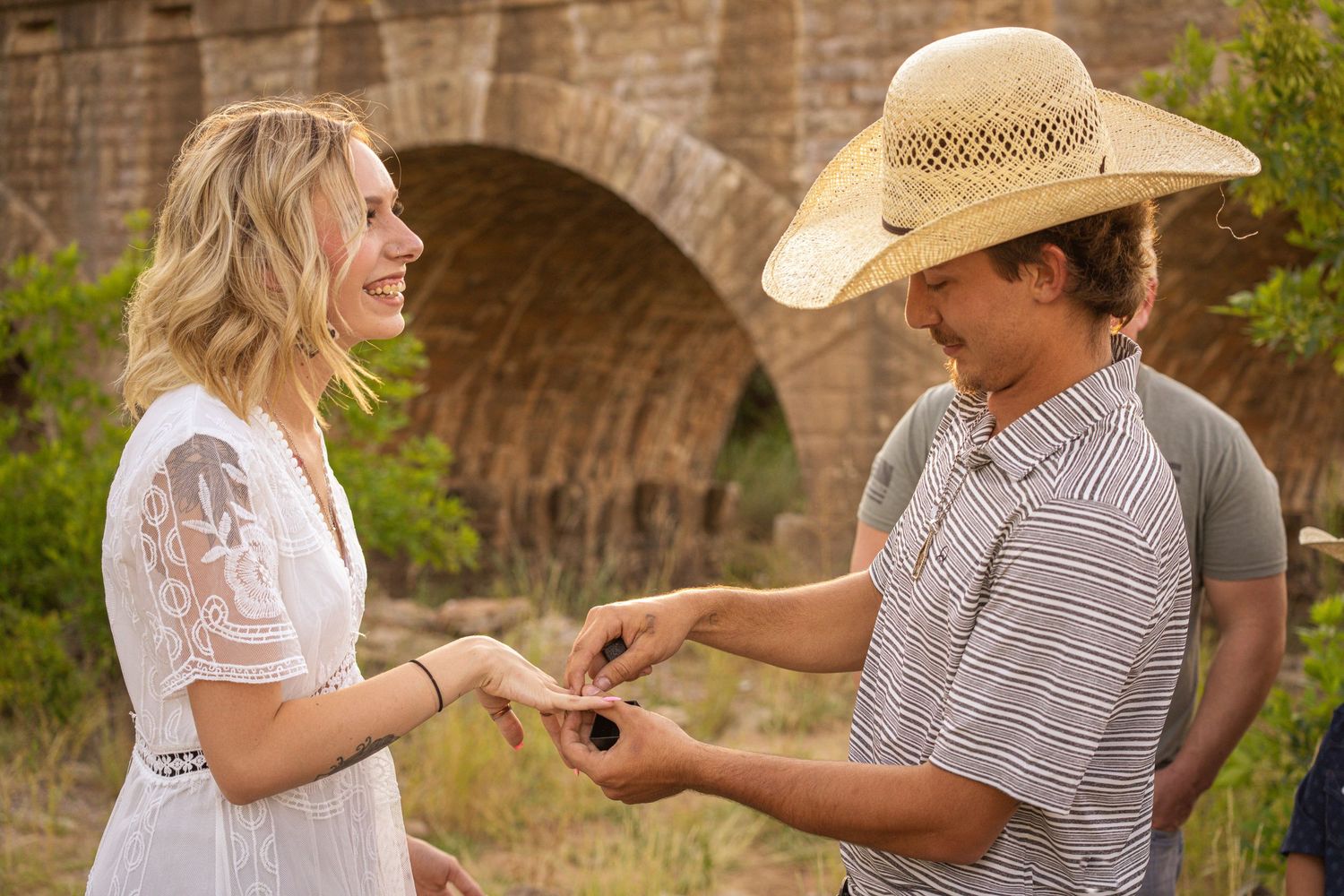 A couple shares a sweet proposal moment under a stone bridge in a natural outdoor setting.